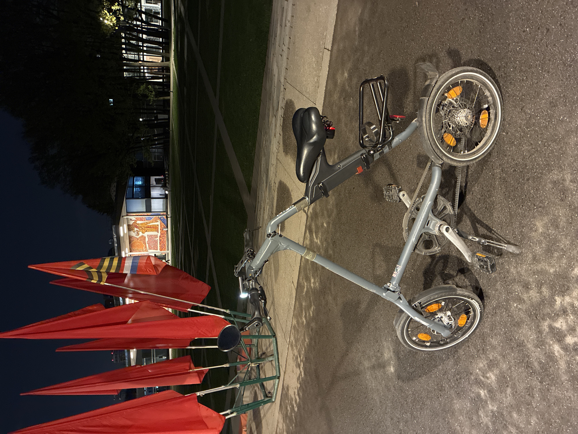 The image shows a small, gray folding bicycle parked on a paved surface next to a grassy area. The bicycle has a black seat, a rear rack, and orange reflectors on the wheels. It appears to be in good condition. In the background, there's a building with a mosaic on its exterior, and several red flags or banners are displayed on metal poles. The time appears to be late evening or night, given the darkness and artificial lighting illuminating the area. The location is identified as Unknown, Unknown.