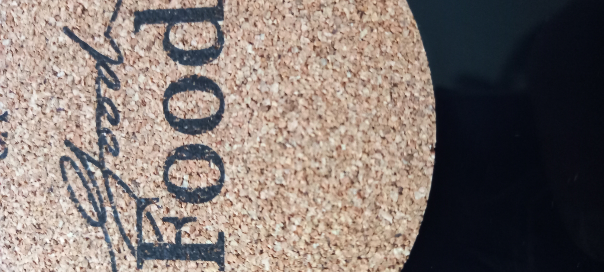 A vertical, close-up photograph captures a portion of a circular cork coaster resting on a flat, uniformly black surface. The coaster is composed of compressed, granulated cork, exhibiting a speckled texture with varying shades of light brown and beige. Black text is printed on the cork surface. The upper word, rendered in a flowing script font, reads "Good". Below it, in a bold, sans-serif block font, the word "Food" is clearly visible. Approximately two-thirds of the coaster's circular form is present within the frame, with the bottom arc disappearing off the lower edge. The black surface beneath the coaster appears to be smooth and minimally reflective. No individuals, actions, or specific environmental details beyond the immediate objects are discernible.
