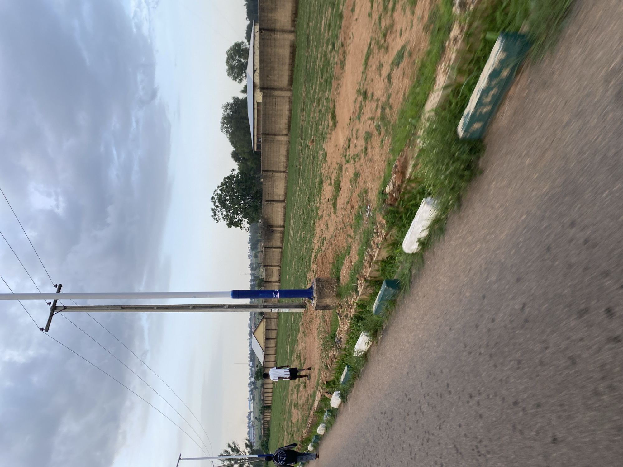 This is a medium-distance, eye-level, outdoor shot of a dirt road in Barikin Jirga Summa, Nigeria. The road is on the left side of the frame and is paved with asphalt. On the right side of the road, there is a low curb with white and green painted concrete blocks. Beyond the curb is a grassy area, followed by a brown dirt field. A concrete wall runs along the far edge of the field, and beyond that are buildings and trees. In the background, there are power lines and utility poles. Two individuals are walking away from the camera on the dirt path next to the road. The sky is overcast with gray clouds.