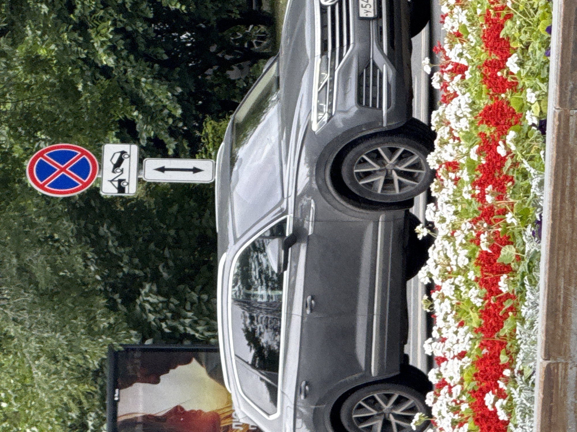 A dark grey Volkswagen Touareg is parked on a city street in Moscow, Russia, as indicated by the license plate starting with "Y54". The car is positioned behind a colorful flowerbed filled with red and white flowers. To the left of the car, a billboard featuring a close-up profile of a woman and a man is partially visible. Behind the billboard and the car, a dense green leafy tree line is present. To the right of the billboard, a street sign with a red circle and a blue cross indicates no stopping, accompanied by a smaller sign depicting a tow truck, and a directional arrow pointing downwards. The sky is overcast.
