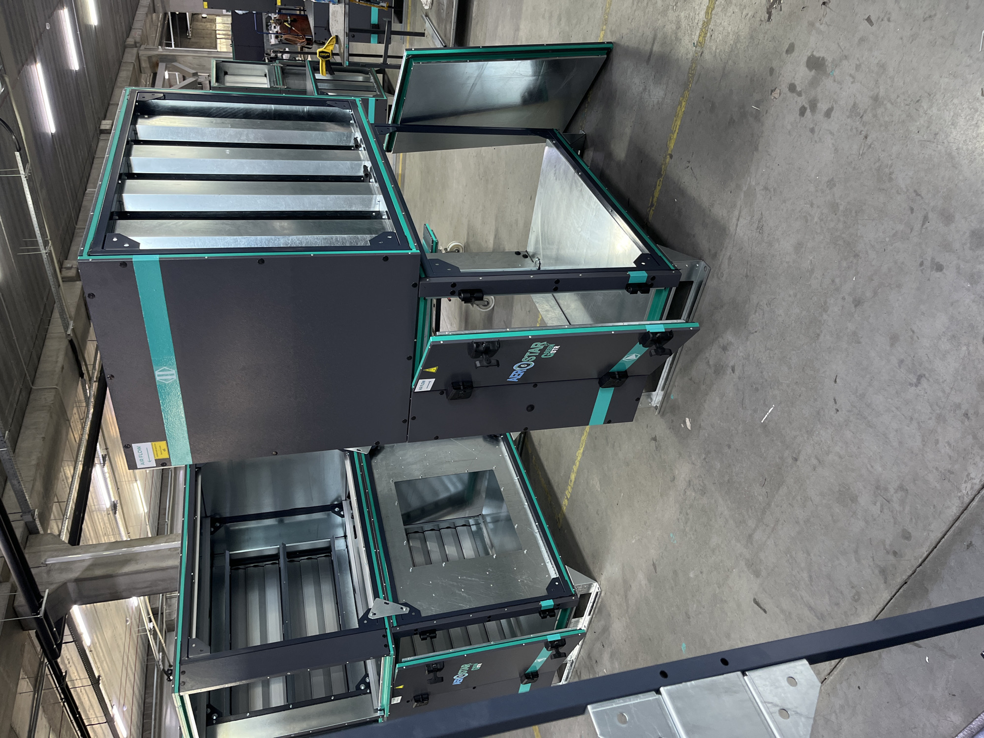 In a large industrial warehouse in Kyiv, Ukraine, two large modular air handling units are positioned on a concrete floor. The units are dark gray with teal accents. The unit in the foreground is partially disassembled, with one side panel open, revealing its internal metal structure. The other unit, partially visible behind it, is similarly constructed. The environment is typical of a manufacturing or storage facility, with exposed ceilings, ductwork, and lighting fixtures. The floor is concrete, marked with yellow lines. No people are visible in the image.