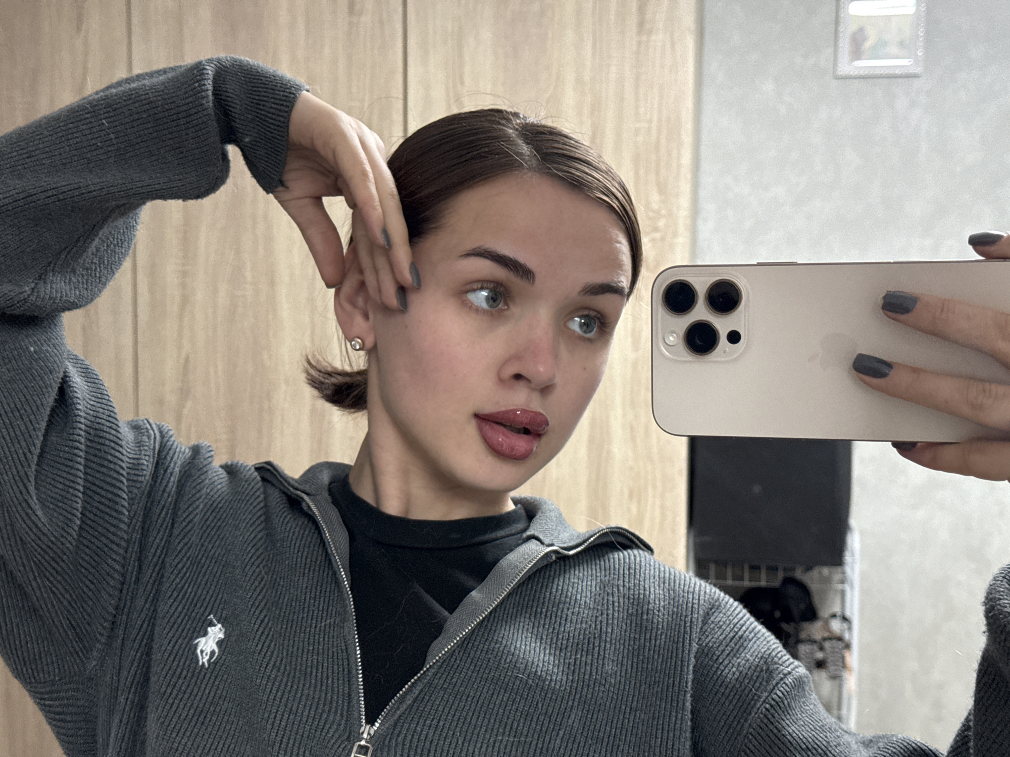 A young woman is depicted in a mirror selfie, holding a light-colored smartphone with a triple camera array in her right hand. Her nails are painted a dark grey. She has dark brown hair pulled back, light-colored eyes, and is wearing a dark grey ribbed zip-up sweater over a black top. The sweater features a small white Ralph Lauren polo player logo on the left chest. Her lips are glossy and slightly parted in a pensive or posed expression, with her gaze directed to her right. Her left hand is raised, with fingers partially covering her left ear, which sports a small silver stud earring.
The background of the mirror selfie suggests an indoor setting, possibly a room with light-toned wooden cabinets or wall panels on the left side and a lighter, reflective wall on the right. A small, decorative square mirror or frame is visible high on the right wall in the reflection. The overall lighting appears to be natural indoor light. The activity is a casual mirror selfie, likely taken in a domestic setting in Лобаново, Russia.