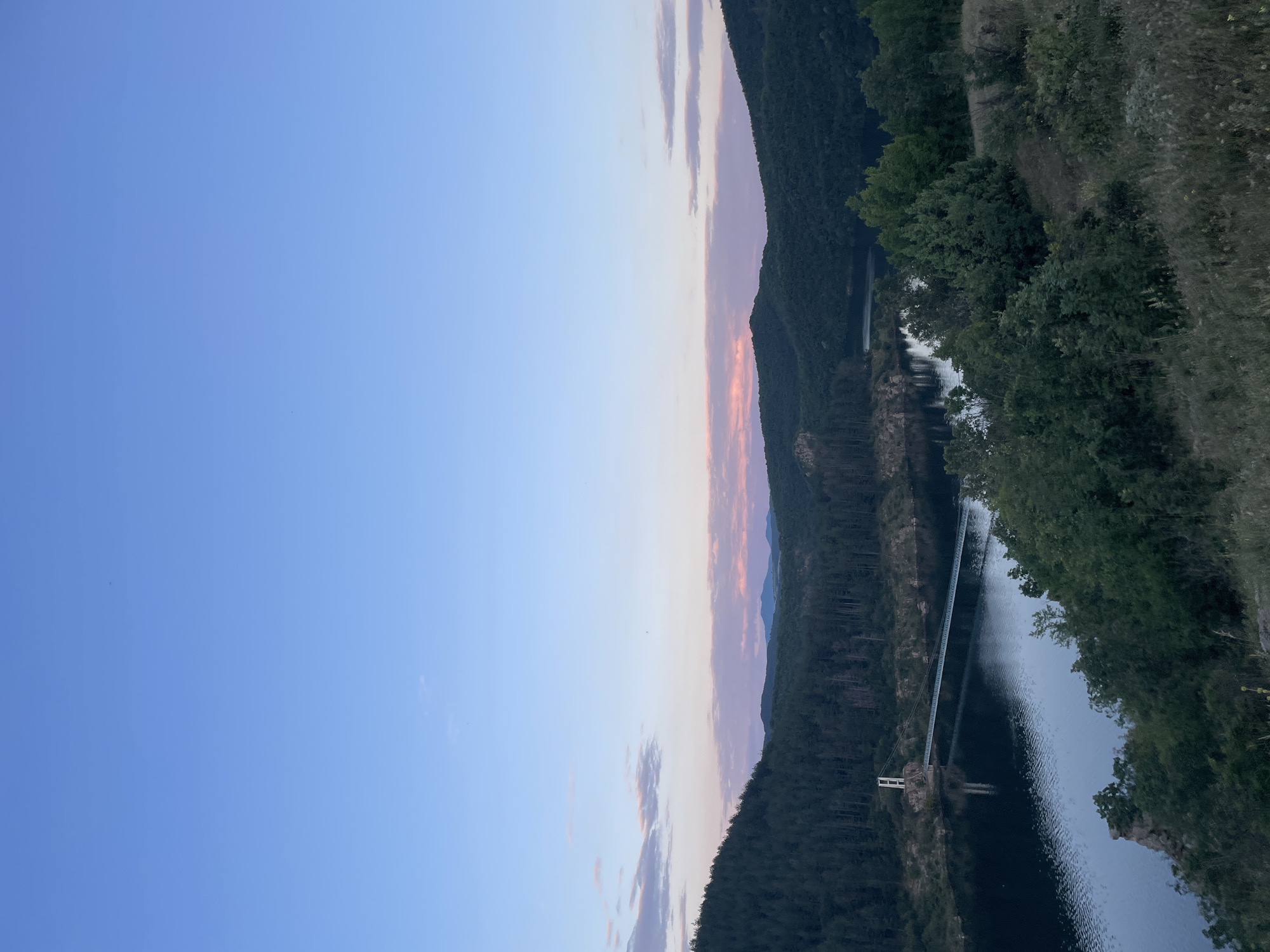 A high-angle, wide shot captures a serene landscape at dusk. The sky transitions from a clear, light blue at the top to a soft gradient of pink and purple hues near the horizon, with a few wispy clouds scattered throughout. Below the sky, rolling, tree-covered hills dominate the midground and background, their dark green foliage creating a textured silhouette against the fading light. A dark, still body of water, likely a lake or reservoir, stretches across the lower portion of the image, reflecting the dusky sky and the surrounding trees. A slender suspension bridge, with a white pylon structure at one end, crosses the water, its reflection shimmering on the surface. The foreground is a hillside covered in dry grass and scattered green bushes, suggesting a natural, undeveloped area. The overall scene evokes a sense of peace and natural beauty. The location is the Yastrebino Reservoir near the village of Yastrebino, in the municipality of Suvorovo, Varna Province, Bulgaria.