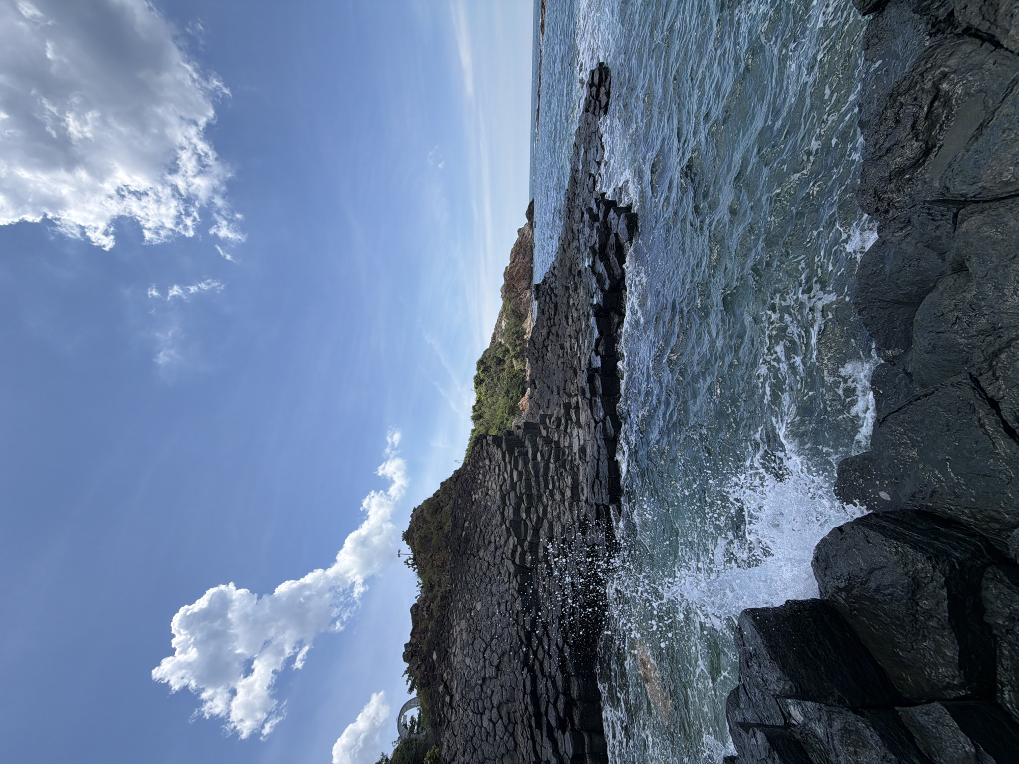 This is a photograph of the Gành Đá Đĩa (Plate-like rocks) in An Ninh Đông Commune, Phú Lương District, Tuy An, Phú Yên Province, Vietnam. The image depicts a dramatic coastline characterized by hexagonal basalt columns that appear to be stacked like discs. These natural formations extend from the land into the turquoise sea, where waves crash against them, creating white spray. The sky above is a bright blue with scattered white clouds. The foreground is dominated by the dark, angular basalt rocks, some of which are wet from the sea. In the middle ground, more of the hexagonal rock formations form a layered cliff. The background shows a lush green hillside with vegetation, and a distant arch-like structure is visible on the left side of the frame. The overall scene is one of natural geological wonder and the power of the ocean. There are no people visible in the image.
