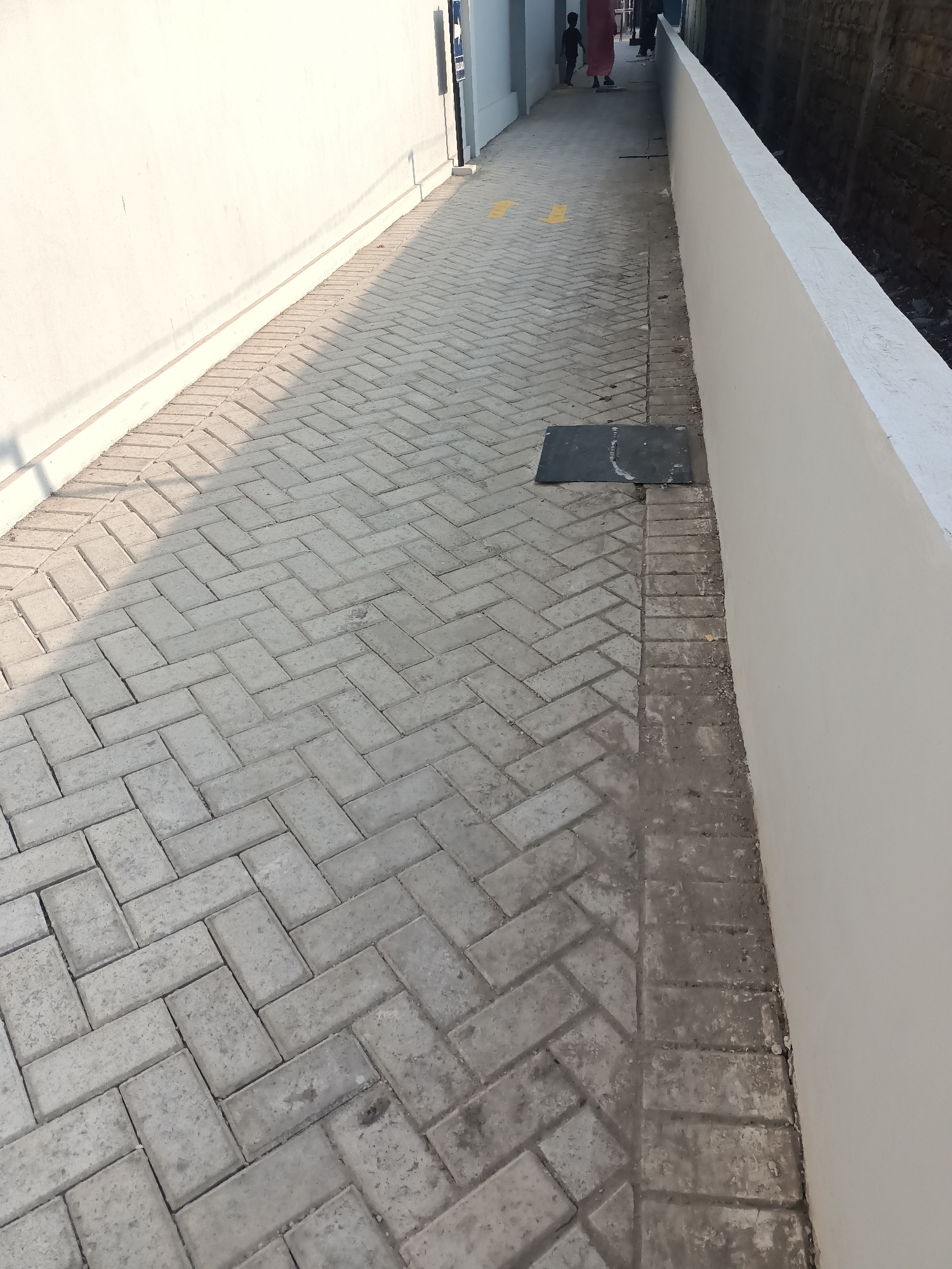 The image depicts a narrow, paved walkway in Purwakarta, Indonesia. The walkway is constructed with herringbone pattern brick pavers. On the left, a smooth, white wall runs along the walkway, casting a long shadow. On the right, a low, off-white colored wall borders the walkway, with a dark, rough wall visible beyond it. Two individuals, one adult and one child, are visible in the distance at the end of the walkway, facing away from the camera and walking towards a building. Yellow arrows are painted on the pavers, indicating direction. A dark, rectangular grate is set into the pavement near the off-white wall. The scene appears to be outdoors in daylight, with the sun casting distinct shadows.