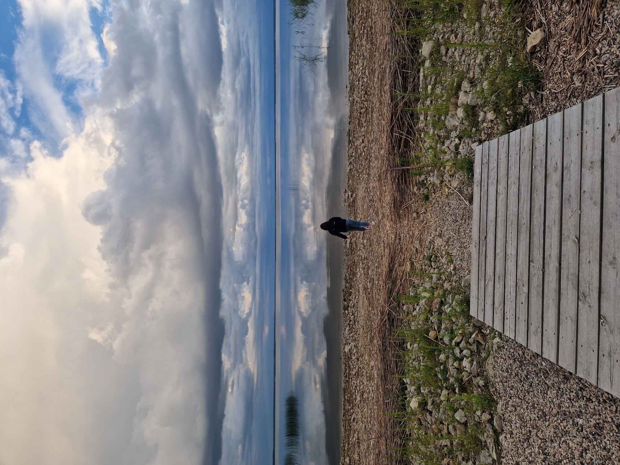 A person wearing a dark hoodie and light-colored pants walks away from the viewer towards a calm body of water. They are walking on a wooden boardwalk that transitions into a gravelly shore. The water's surface is highly reflective, mirroring the dramatic, cloudy sky above. Tall reeds or grass are visible along the water's edge and in the distance. The scene appears to be a natural environment, possibly a lake or large pond, on the outskirts of Īdeņa, Latvia. The overall mood is serene and contemplative, with the vast sky and reflective water dominating the composition.