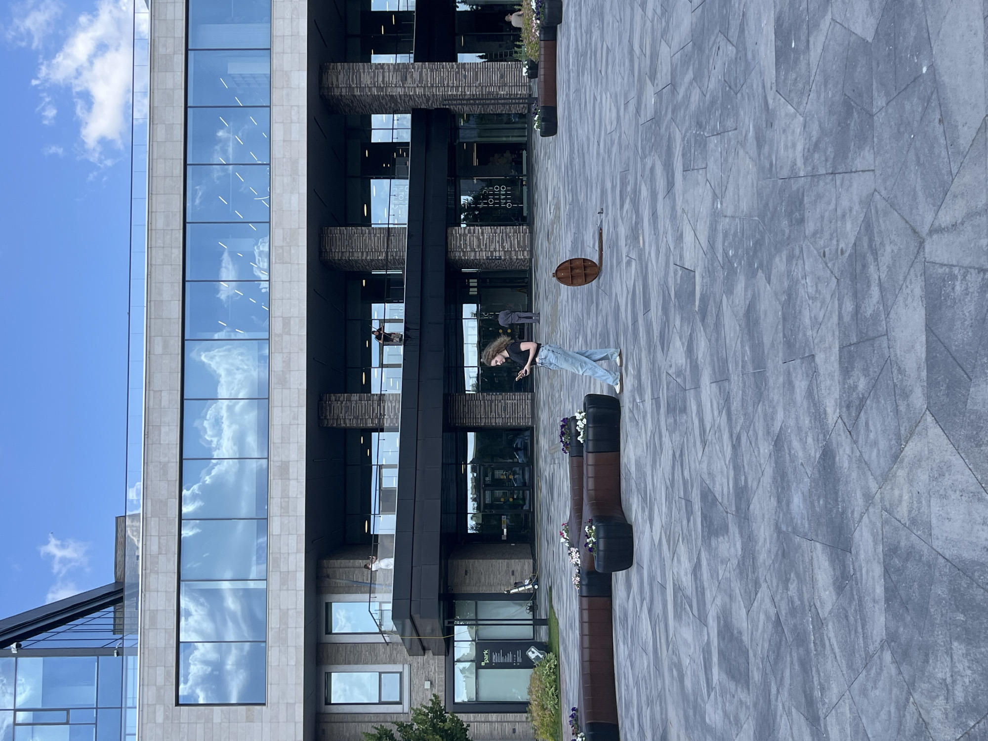 A woman is standing in an open, paved plaza in front of a modern building with large glass windows. She is wearing a black t-shirt and light-wash jeans and is looking at her phone. In the background, there are several people visible on a balcony and inside the building. The building appears to be made of stone and glass, with dark columns supporting an upper level. There are benches with planters and flowers in the plaza. The sky is blue with some clouds. The image appears to be taken during the day. The location of the scene is Kazan, Russia, specifically the IT Park named after Bashir Rameev.