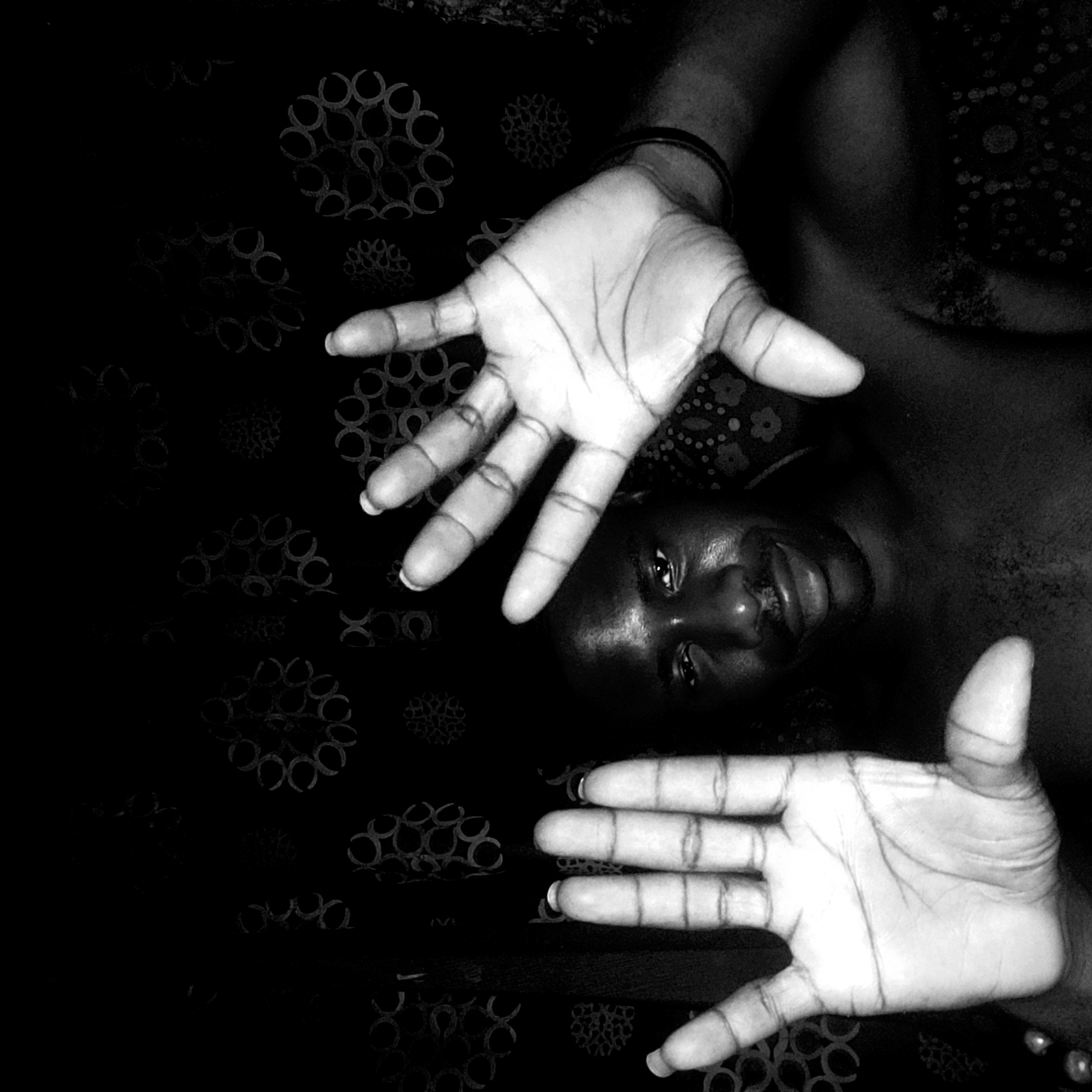 A black and white photograph shows a person lying down, seemingly on a bed or patterned surface. The person's face is visible, positioned near the center of the frame, with their eyes open and a slight smile. Two hands are prominently displayed in the foreground, appearing to reach towards the viewer. The background consists of a dark, patterned fabric. Given the prompt's requirement of including Narayi, Nigeria, though without explicit confirmation in the image, we infer that this scene potentially represents someone lying on a bed in a residential location in Narayi, Nigeria.