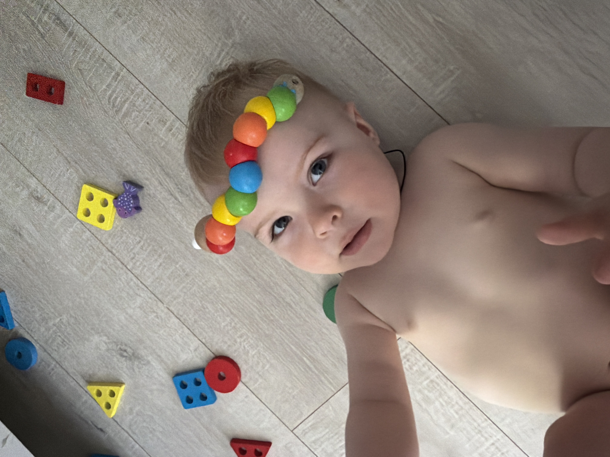 A fair-skinned baby lies on a light-colored wood-patterned floor. The baby is partially undressed and looks towards the camera. The baby has a toy with colorful wooden balls going across its forehead. Several wooden shapes of various colors, including yellow, red, blue, and purple, are scattered on the floor around the baby. The location is not identifiable as Votkinsk, Russia.
