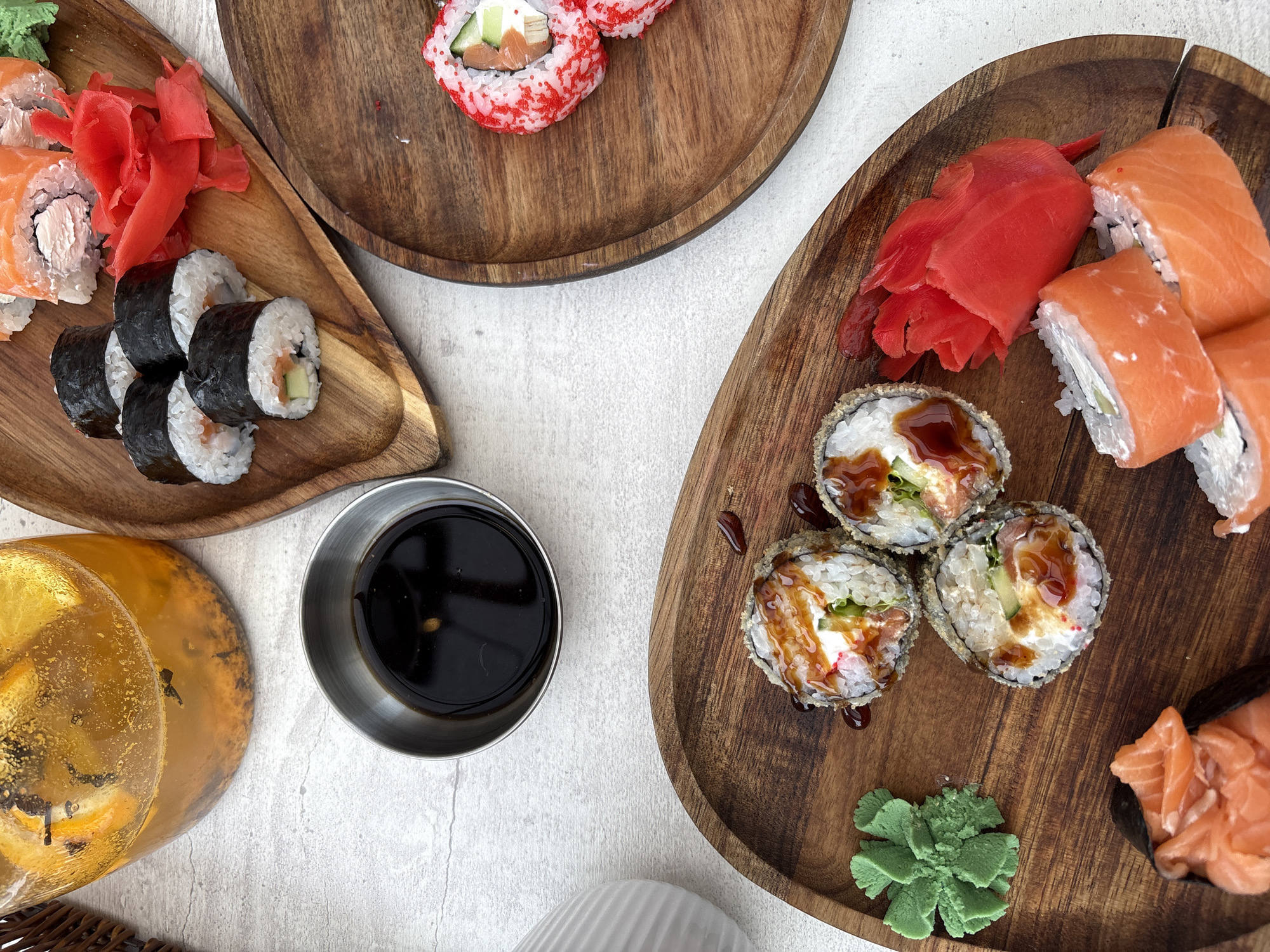 An overhead, close-up shot shows a variety of sushi and a pitcher of infused water arranged on a light-colored textured surface. In the upper left, a glass pitcher contains a golden liquid with visible slices of orange and dark specks. To the right of the pitcher, a small, silver bowl holds dark soy sauce.
A wooden platter in the upper middle of the frame displays several pieces of sushi wrapped in dark seaweed, with visible white rice and orange salmon. Beside the sushi are several bright pink pickled ginger slices.
Another wooden platter, positioned to the right, holds a single piece of sushi with red roe on the outside, featuring salmon and cream cheese.
A third wooden platter occupies the bottom of the frame, showcasing a selection of sushi. Three pieces in the center appear to be fried or tempura rolls, drizzled with a dark sauce, and are accompanied by a bright green mound of wasabi. To the left of the wasabi, a piece of nigiri sushi with salmon is visible. At the bottom right of this platter, several pieces of salmon rolls are arranged.
The overall scene suggests a meal or gathering focused on sushi and beverages. The environment is a flat, textured surface, likely a tabletop, in a well-lit indoor setting. The image lacks any identifiable landmarks or elements that would suggest a specific location in Ust-Ilimsk, Russia.