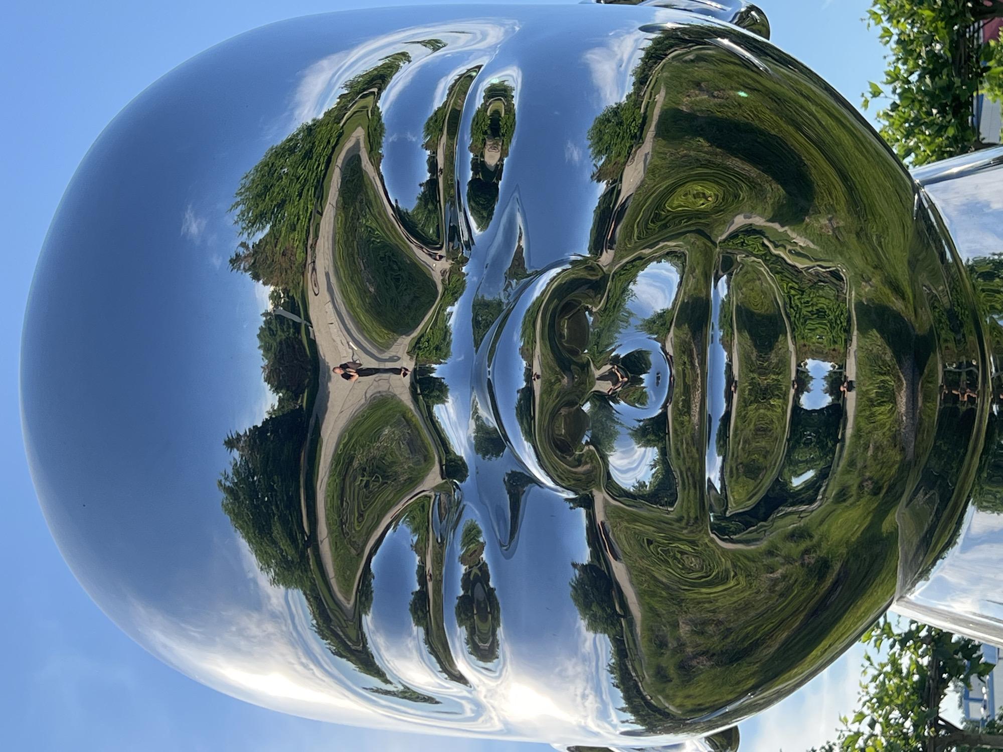 This is a close-up, reflective shot of a large, silver sculpture of a baby's head, set against a bright blue sky. The sculpture is highly polished, creating distorted reflections of its surroundings. In the reflections, a pathway winds through lush green grass and trees. Several people are visible as small figures within the reflections, including one person standing near the pathway who appears to be taking a photograph. The sculpture itself is smooth and featureless, with the reflections creating the impression of eyes, nose, and mouth. The overall impression is one of surrealism and playful distortion, highlighting the reflective properties of the artwork and its interaction with the environment. The sculpture is located in the city of Krasnodar, Russia.
