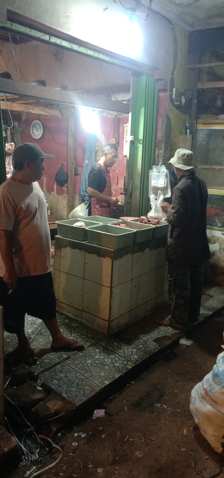 A scene at a meat stall in the city of Subang, Indonesia. Three men are present. One man wearing a light-colored t-shirt and a baseball cap stands to the left, facing the center of the stall. Another man, wearing an apron over a dark shirt and black pants, is positioned behind the counter, appearing to work with meat. A third man, in a dark jacket and a light-colored hat, stands to the right, facing the man behind the counter. The stall features several green plastic containers filled with raw meat, arranged on a tiled counter. The ground in front of the stall is wet and tiled, with debris and some plastic packaging visible. The lighting appears to be artificial, casting shadows and illuminating the scene. The background suggests an indoor market or covered area, with visible pipes and structural elements. A clock is mounted on the wall.