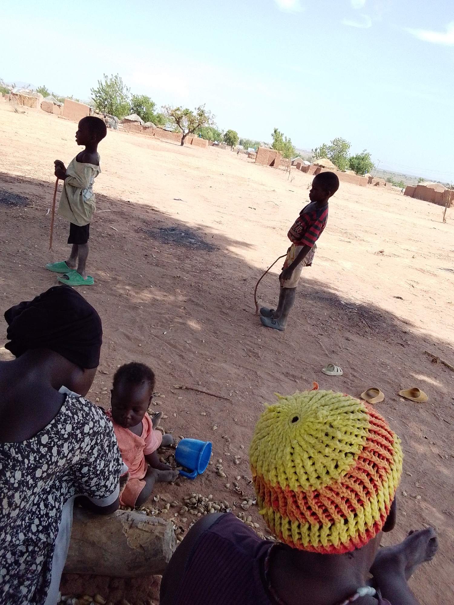 The image shows a scene in Mulke Barikin Sayejo, Nigeria. Two children stand in a dusty, open area with sparse vegetation and scattered buildings in the background. One child wears a light-colored top and dark shorts, holding a stick. The other wears a striped shirt and light shorts, holding what appears to be a looped stick. In the foreground, an adult wearing a black head covering sits near a log, a baby sits on the ground nearby playing with a blue cup. Another person wearing a colorful yellow and orange crocheted hat sits with their back to the camera. Scattered peanuts are on the ground near the people sitting. Several pairs of sandals are scattered on the ground.