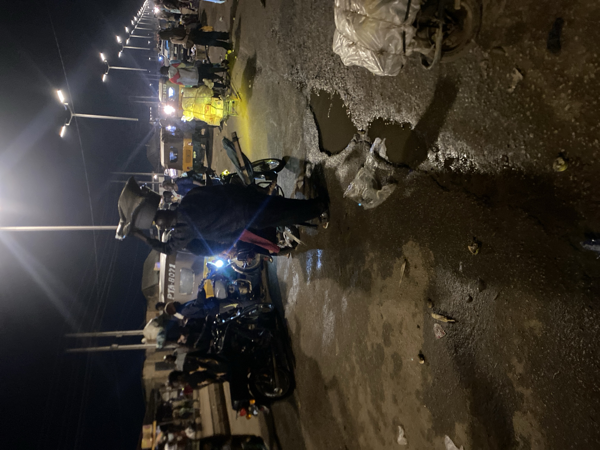 This image captures a bustling street scene at night in Bukuru, Nigeria, illuminated by numerous streetlights and vehicle headlights. The ground is wet and uneven, featuring prominent potholes, some filled with standing water that reflects the artificial lights.
Numerous people populate the scene, walking, standing, and interacting, suggesting a lively marketplace or transport hub. In the foreground, a person is visible near motorcycles, and further back, others are clustered around vehicles. One person is seen carrying an item on their head.
Vehicles are a significant part of the activity. Several motorcycles, commonly known as *okada*, are present, either parked or in motion. Multiple yellow auto-rickshaws, or *keke napeps*, are visible, some laden with goods in large, clear plastic bags. A larger white vehicle, possibly a small bus or truck, is also in the mid-ground.
Notable details include the extensive line of modern streetlights stretching into the distance, casting bright, stark illumination across the dark environment. The road surface is damaged, with large, deep potholes affecting vehicle and pedestrian movement. Various pieces of litter, including plastic bags and other debris, are scattered on the wet ground. The overall atmosphere is one of active nighttime commerce and transit.
Visible text includes "PTA-8071" on the side of the white vehicle and the partial word "SAFE" printed on the fabric cover of one of the yellow auto-rickshaws.