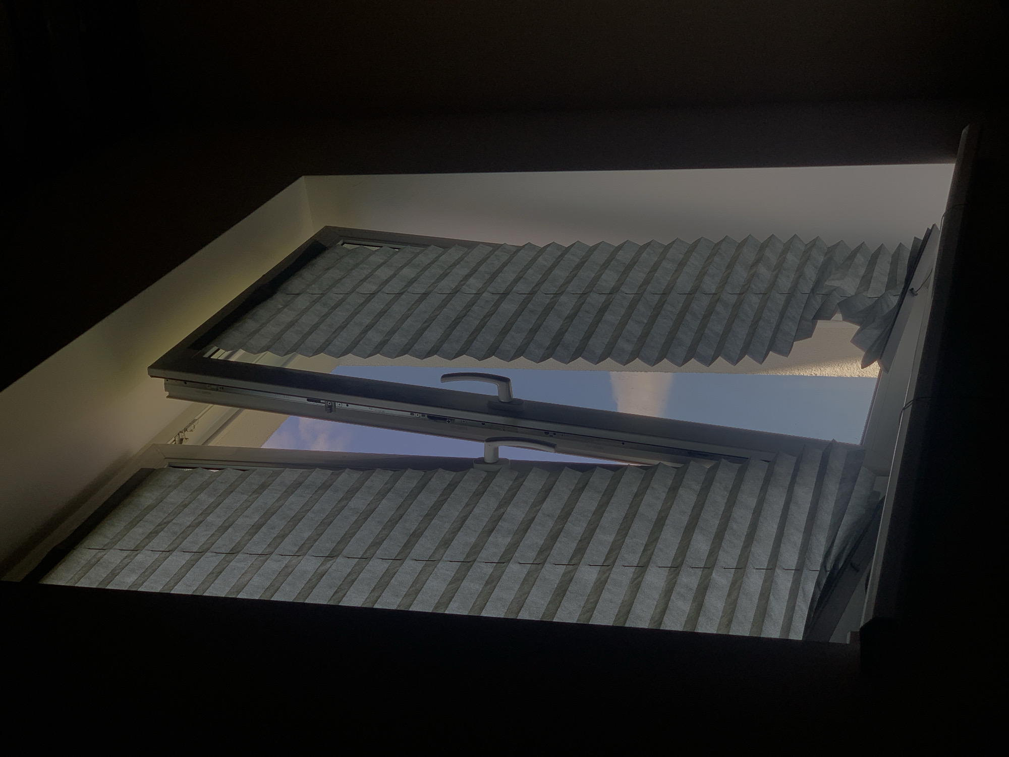 The image shows an open window with pleated blinds. The window frame is white, and the blinds are gray. The left blind is mostly closed, while the right blind is open and pulled to the side, revealing a blue sky with some clouds. The blinds appear to be slightly damaged, with some pleats detached on the right side. The surrounding environment is dark, suggesting the photo was taken indoors, possibly at dusk or dawn. There is no indication of the specific location within the city of Minsk, Belarus, or any people or interactions.
