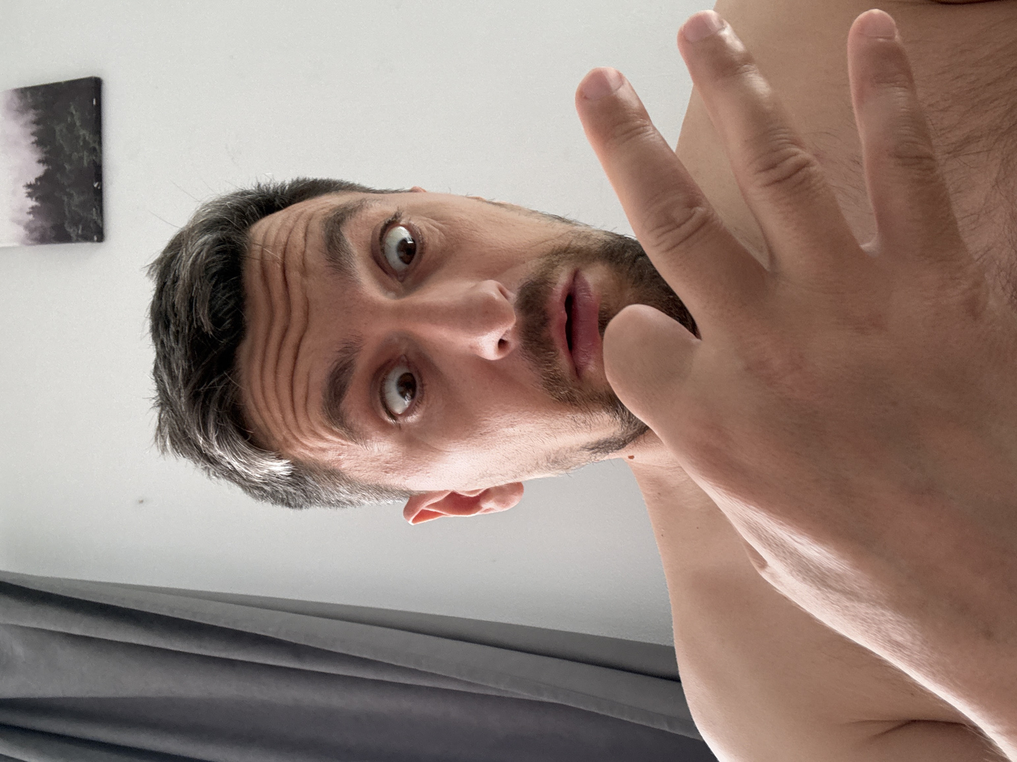 A close-up, upward-angled view of a light-skinned male with graying, short hair and a dark beard. The man's facial expression is surprised, and his mouth is slightly open. One hand is raised, palm outward, obscuring part of his face. The background consists of a white ceiling, a gray curtain, and a black-and-white landscape picture hanging on the wall. The scene is indoors. The location is Unknown, Unknown.