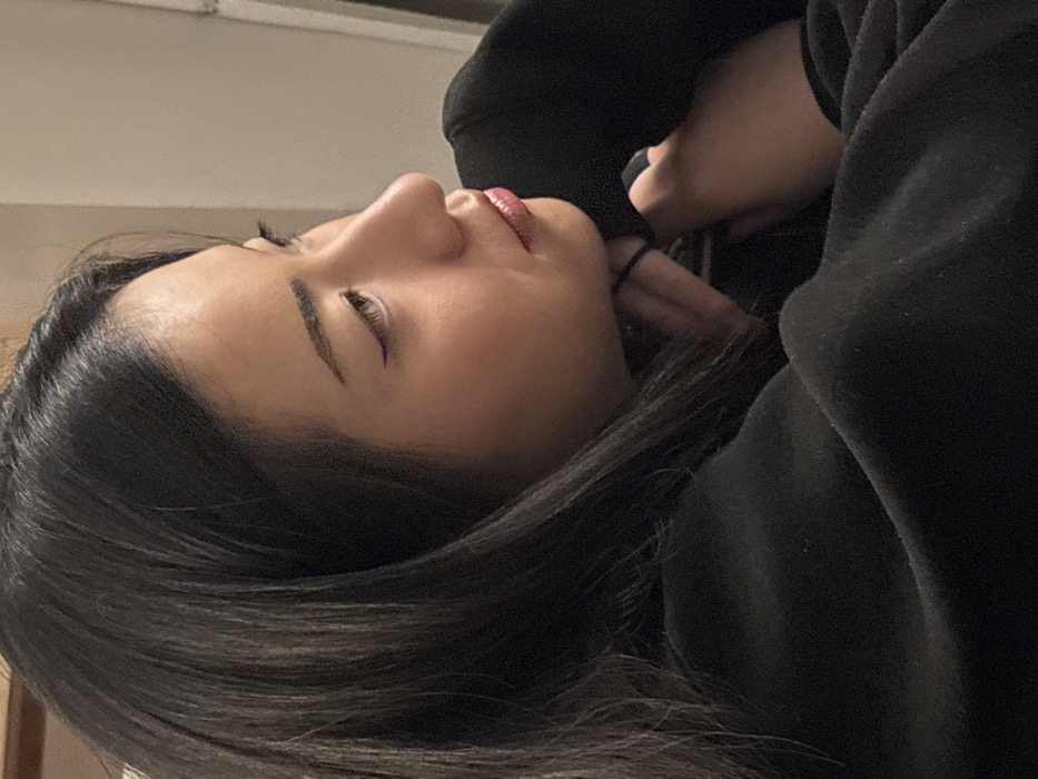 The image depicts a close-up, profile view of a young individual, positioned facing towards the right side of the frame. The subject has dark, straight hair styled with a side part, falling forward over the shoulders. Her visible eye shows winged eyeliner, and her lips are subtly colored. She is wearing a dark, long-sleeved garment, possibly a sweatshirt. Her right hand is raised, with fingers supporting her chin or jawline. A black elastic hair tie is present on her right wrist or wrapped around her fingers. The background is out of focus and appears to be an interior space with light-colored walls, indicating an indoor setting. There are no identifiable landmarks or features within the image to specify a location in Paris, France.
