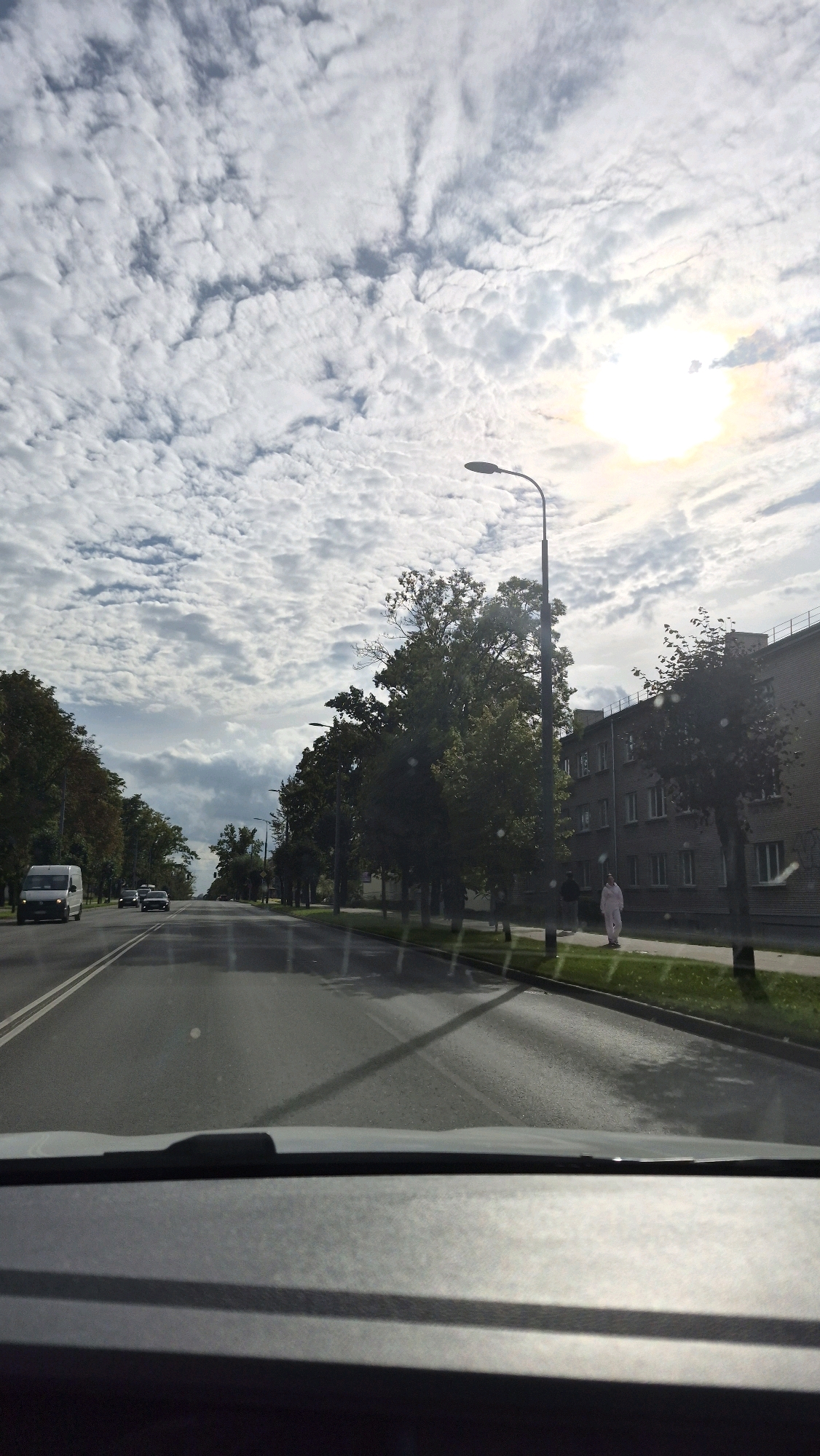 The image, taken from inside a vehicle, depicts a street scene in the city of Rezekne, Latvia. The road is paved with asphalt and has two lanes in each direction, separated by white lines. Trees line the right side of the street, with a building behind them. On the left side of the street, more trees are visible, and a white van is parked. The sky is filled with scattered clouds, and the sun is shining brightly, casting shadows on the road. A lamppost stands on the right side of the road, and a person in light-colored clothing is walking on the sidewalk. The overall impression is of a typical urban environment on a partly cloudy day.