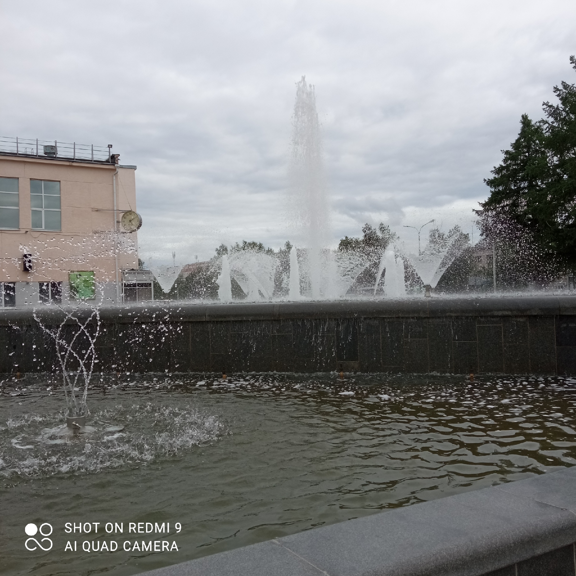 The image depicts a fountain in the city of Chaikovsky, Russia. The fountain is constructed of dark stone with a raised edge of gray stone. Multiple jets of water spray upwards and outwards from the fountain's surface, creating a dynamic display. The water cascades back into the pool, creating ripples and reflections on the surface. In the background, a light pink building with windows and a clock on its facade is visible to the left of the fountain. To the right, a tall green coniferous tree stands. The sky is overcast with gray clouds, creating a muted light. The foreground shows the rim of the fountain. A watermark, from the camera used to take the image, is also visible in the bottom left corner.