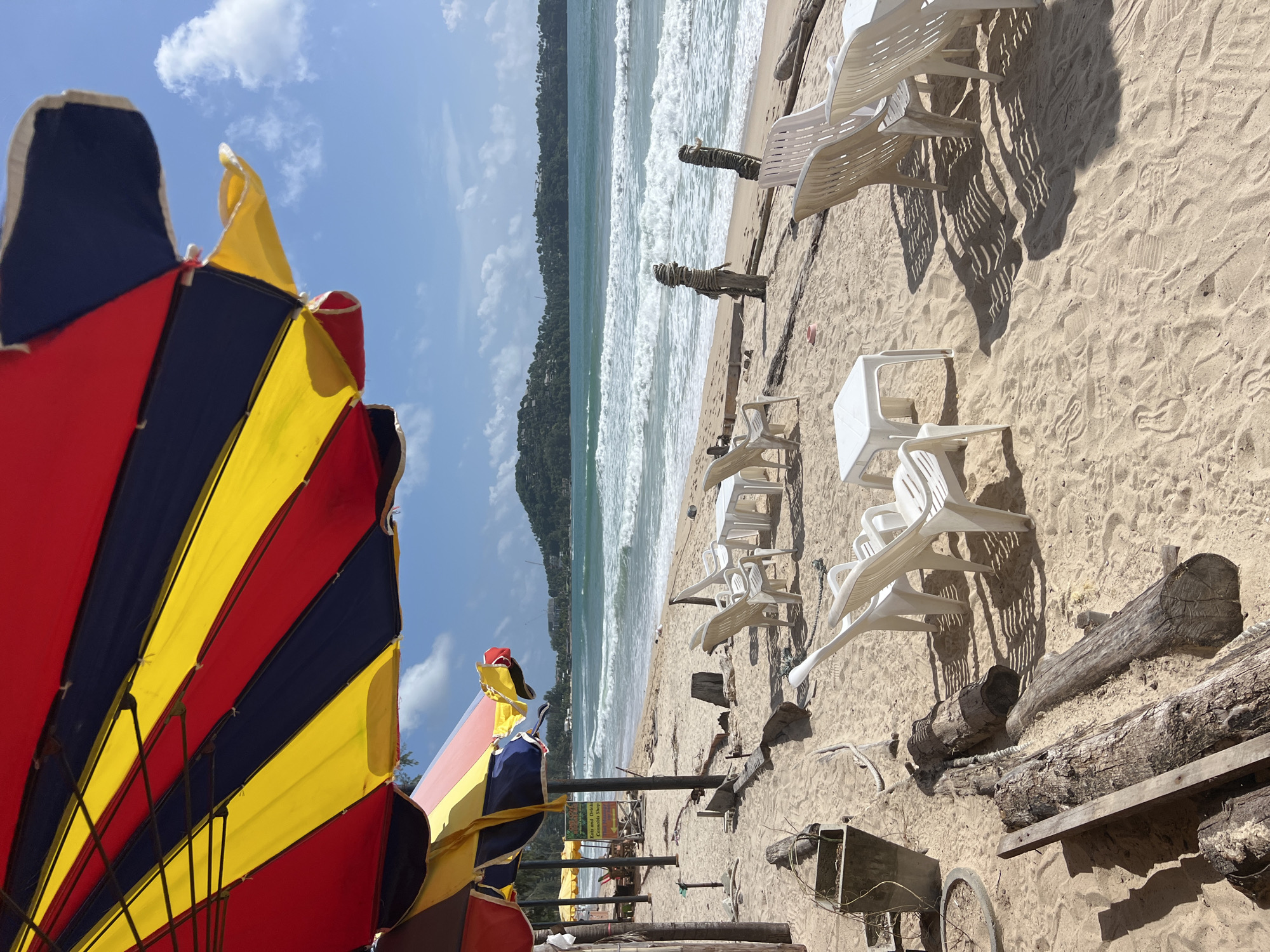 The image depicts a beachfront scene in Choeng Thale, Thailand. A vibrant, multi-colored umbrella, striped in red, yellow, and dark blue, dominates the foreground, casting a shadow on the sandy beach. Scattered across the sand are several white plastic chairs and small white tables, suggesting an outdoor seating area for a beachside establishment. In the middle ground, the ocean rolls in with white-capped waves under a bright, partly cloudy sky. In the distance, a lush, green, hilly coastline is visible. The beach itself is composed of fine, light-colored sand, with various pieces of driftwood and some debris, including a concrete planter and what appears to be a discarded tire rim, visible in the foreground. Two rustic, rope-wrapped wooden posts stand near the water's edge. Further back, more beach umbrellas and a sign can be seen, hinting at commercial activity. The overall impression is a relaxed, tropical beach setting.