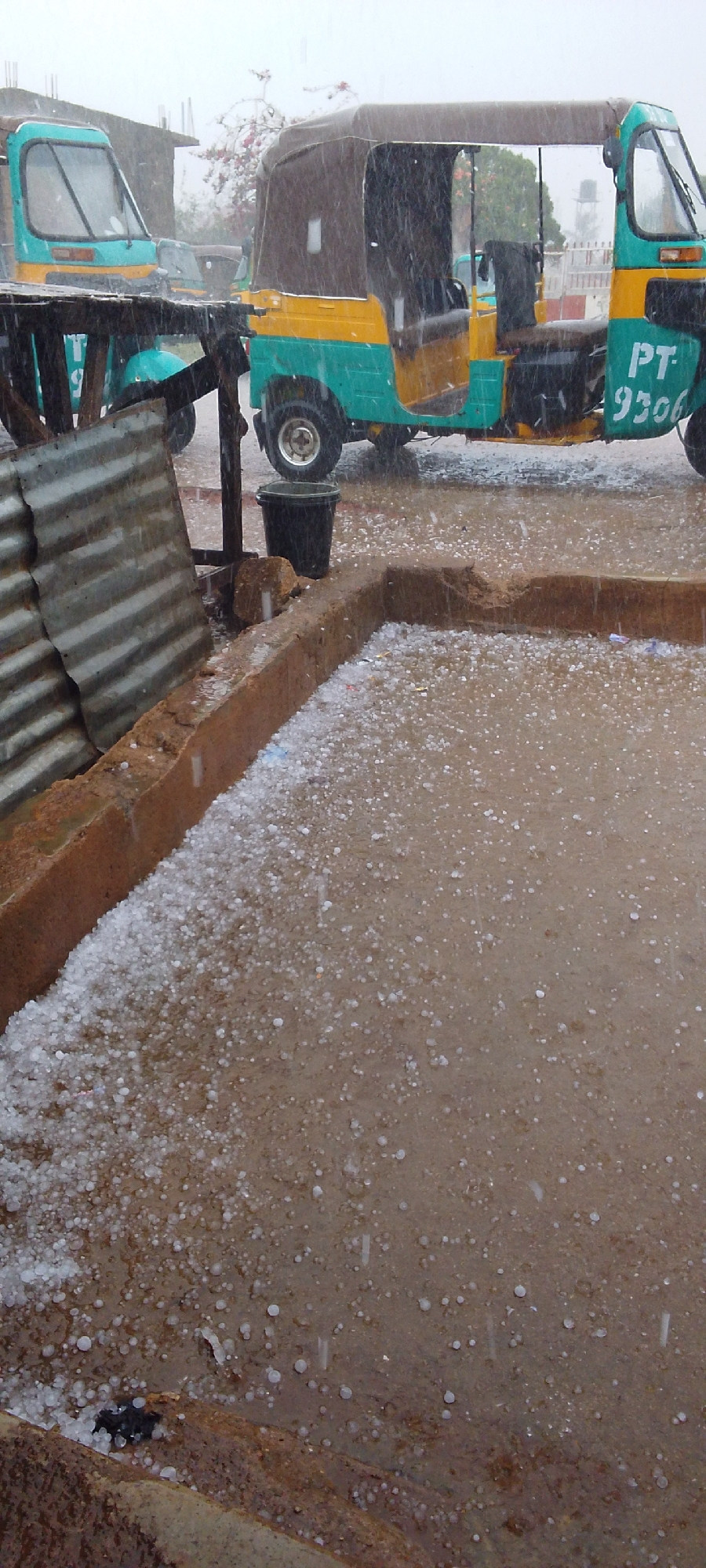 The image depicts a street scene in Jos, Nigeria, during a hailstorm. In the foreground, hail covers a concrete surface, transitioning from a dense accumulation near a low brick wall to a sparser distribution further away. Raindrops are visible amidst the hail. In the background, two three-wheeled vehicles, known locally as "Keke Napep," are parked. One vehicle, partially obscured, is turquoise. The other is turquoise and yellow with a brown canvas roof and the numbers "PT-9306" painted on its side. A black bucket sits on the wall next to the Keke. Corrugated metal sheets are partially visible to the left. The heavy rain and hail reduce visibility.