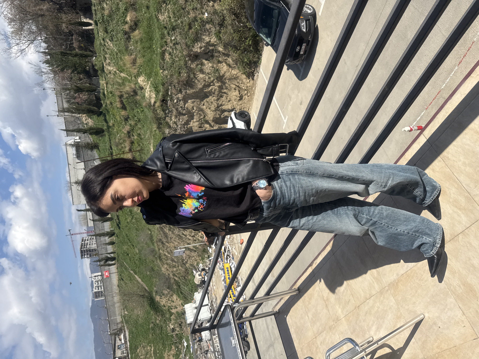 A woman stands on a balcony overlooking the city of Tbilisi, Georgia. She is wearing a black leather jacket over a black t-shirt with a colorful design, blue jeans, and black boots. She also has a watch on her left wrist. Behind her is a black car parked near a railing. In the background, there is a view of Tbilisi, including buildings, cranes, and vegetation. The weather appears sunny with some clouds.