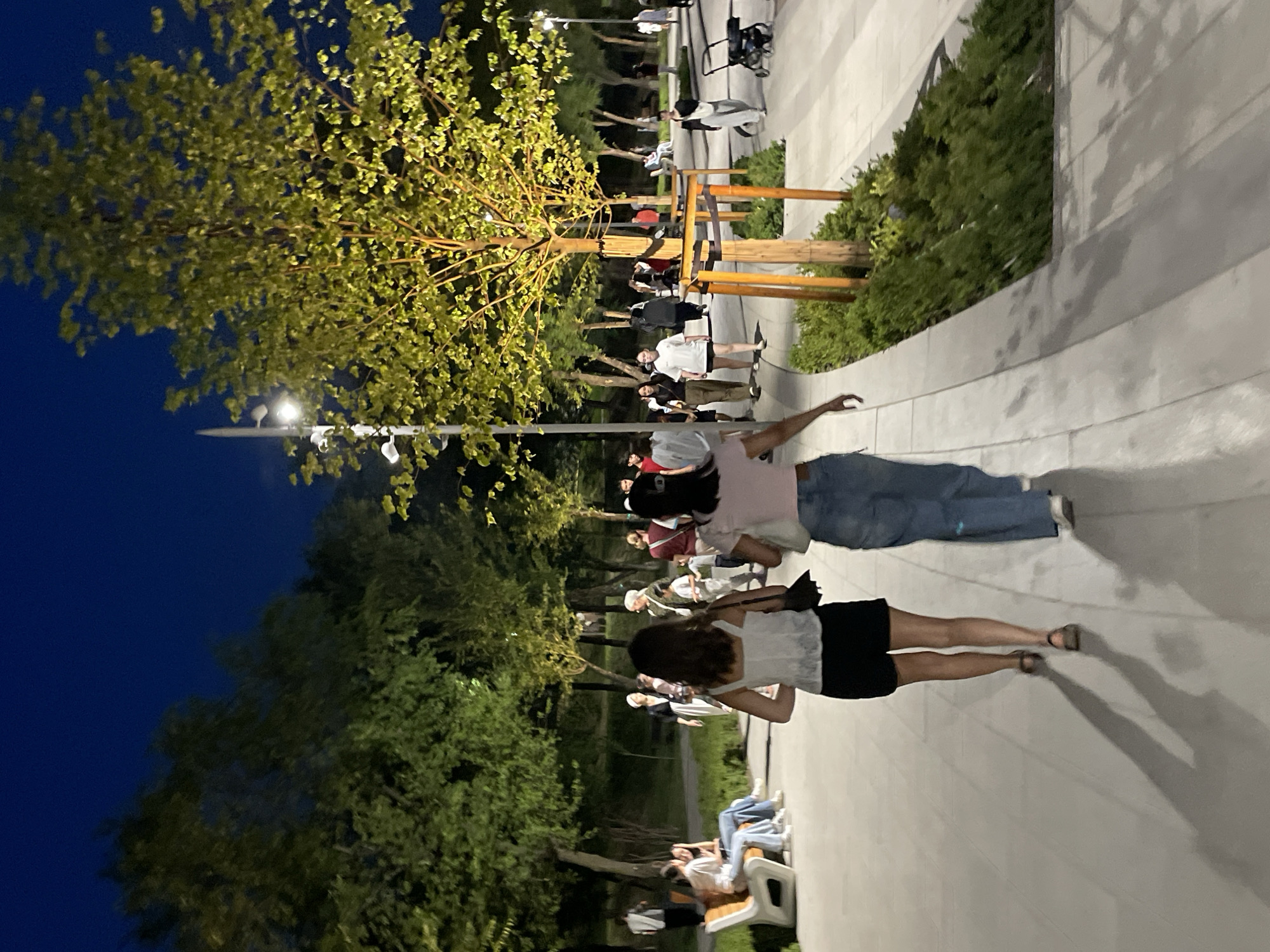 The image depicts a park at night in Almaty, Kazakhstan. The sky is a deep blue, indicating it is dusk or night. Several trees with illuminated foliage are visible, suggesting artificial lighting. A paved pathway winds through the park, with several people walking on it. In the foreground, two young women are walking away from the camera. The woman on the left is wearing a white tank top and black shorts, while the woman on the right is wearing a pink top and blue jeans. Other individuals and groups of people are scattered throughout the park, some sitting on benches and others walking. The park appears to be a public space with benches, lampposts, and well-maintained greenery. The overall scene suggests a relaxed evening atmosphere.
