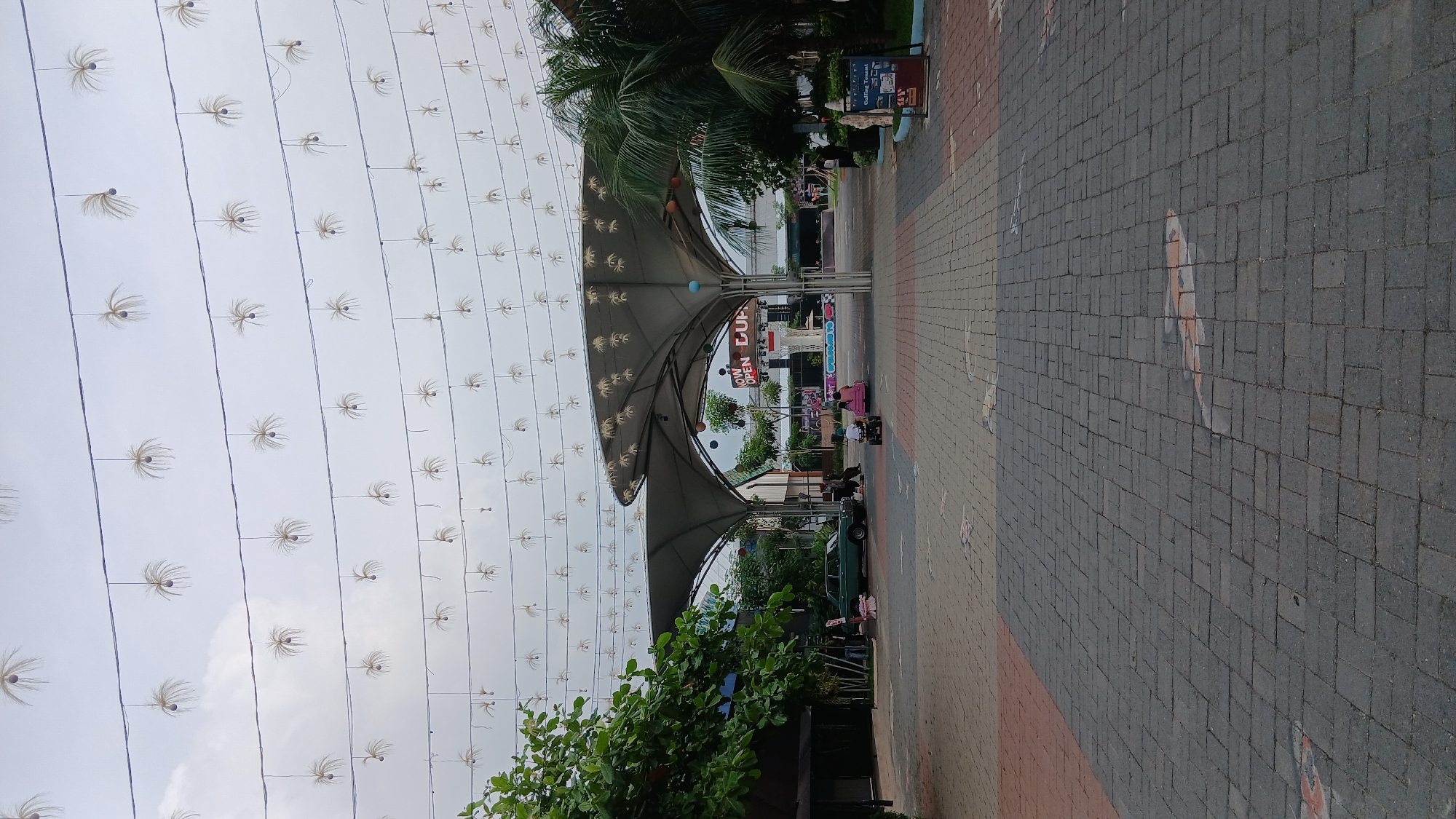 This is a vertical, eye-level shot of an outdoor public space in the City of Medan, Indonesia, likely a park or recreational area, under an overcast sky. The foreground is paved with dark gray interlocking bricks, with scattered patterns resembling stylized fish or koi designs in muted orange and white. Moving further back, the pavement transitions to a mix of reddish-brown and gray bricks.
Dominating the mid-ground is a large, grey, tensile fabric canopy structure with multiple undulating peaks, supported by pillars. Beneath the canopy, a stage or performance area is visible, with a large screen and a sign that reads "NOW OPEN CUP".
To the left of the canopy, a lush green tree with dense foliage extends into the frame, partially obscuring a dark-colored building. Further left, a vintage green sedan is parked.
In front of the canopy, several people are present. A person is sitting on a bench or stool near the parked car. Two children are on a small, red ride-on toy vehicle, with one child steering. Another person appears to be standing near the ride-on toy.
Above the scene, numerous strings of decorative lights are suspended across the sky. These lights are shaped like delicate, spiky, dandelion-like or starburst ornaments, with a small dark orb at their center, hanging from dark wires stretched horizontally.
To the right of the canopy, palm trees are visible, and a small signpost with a red header and white text is placed near the edge of the paved area. The overall impression is of a public gathering or entertainment space, decorated for an event.