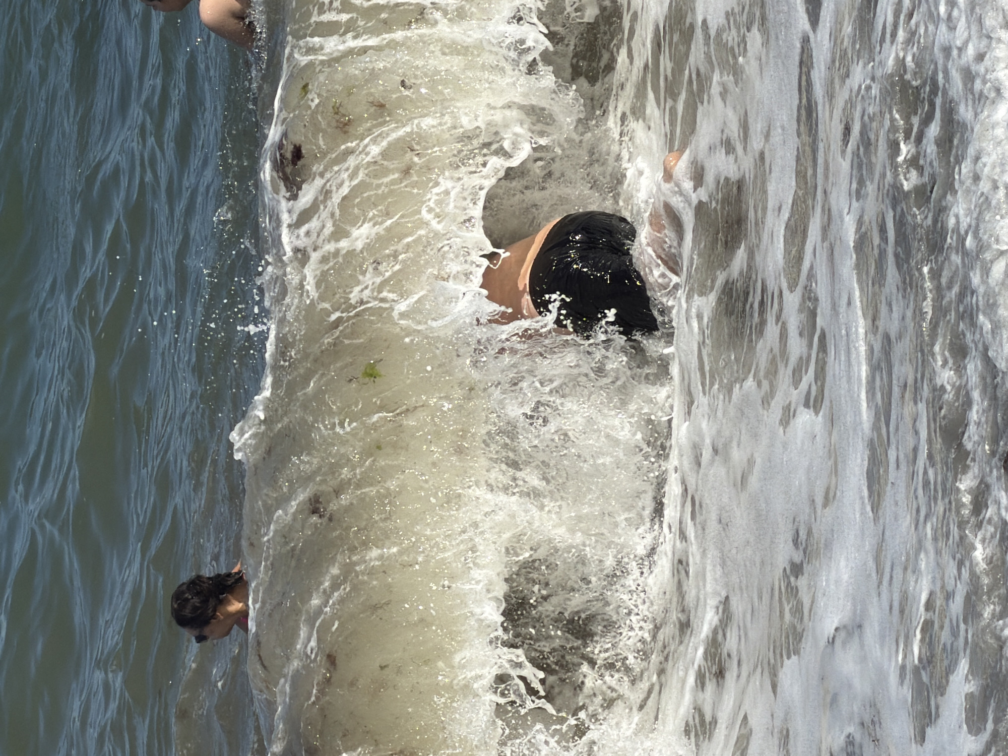 The image shows a person partially submerged in the ocean waves near the shore. The person is wearing black swim trunks and appears to be in the process of being tumbled by the water. The waves are breaking, creating a lot of foam and spray. In the background, another person is visible in the water, and a third person is partially visible at the far right. The scene is likely a beach in Alushta, Ukraine, given the context.