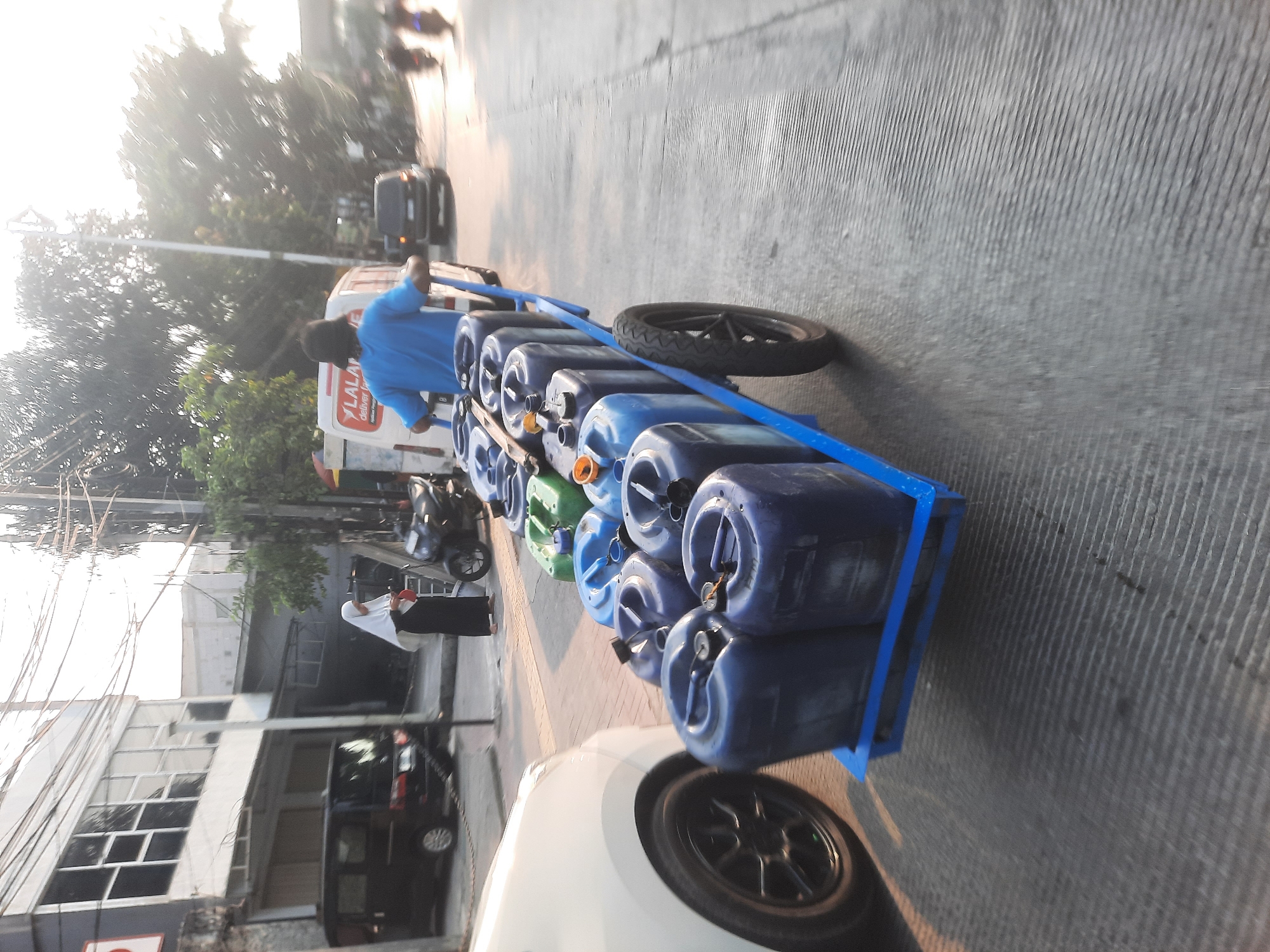 The image captures a scene on a city street in North Jakarta, Indonesia, during what appears to be late afternoon or early morning due to the low sun angle and long shadows. A man wearing a blue shirt is pushing a blue cart laden with numerous blue and green plastic barrels. These barrels are filled with liquid and appear to be of various sizes. The cart has a single wheel at the back. In the background, a white van with a "YALALAL" logo is visible, suggesting it might be a delivery vehicle. Other vehicles, including a black car and motorcycles, are present on the road, indicating urban traffic. A woman in a white hijab and black attire is standing on the sidewalk, looking down. Buildings line the street, and electrical wires are strung overhead. The ground is paved with asphalt, and the overall environment suggests a commercial or residential street in an Indonesian city.