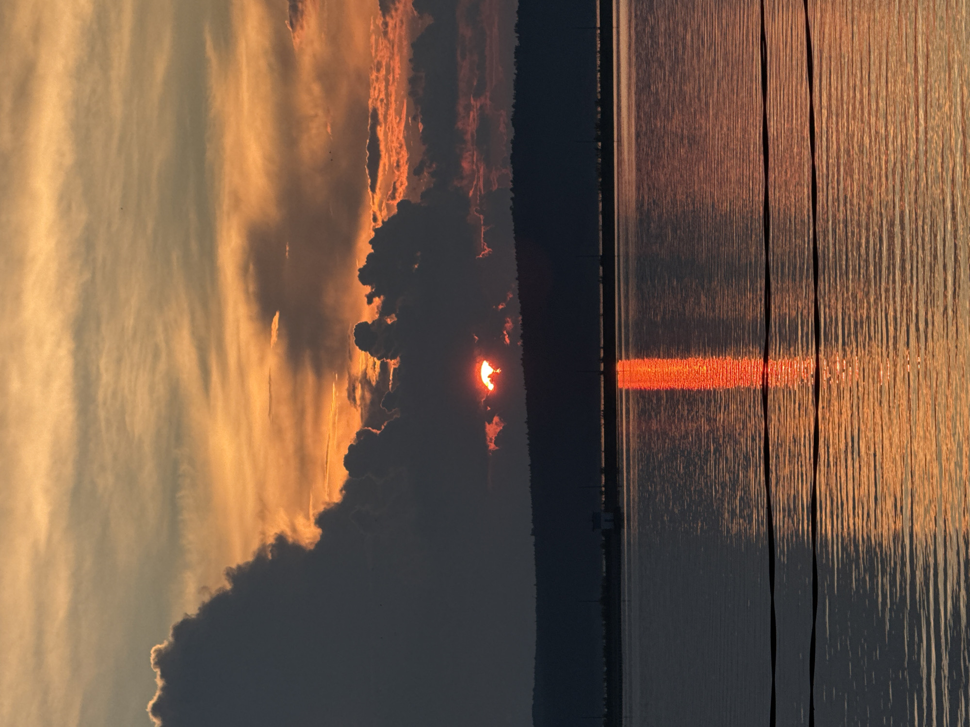 This photograph captures a sunset over the Kama River in Chaikovsky, Russia. The sky is a dramatic display of orange and yellow hues, with large, dark clouds obscuring parts of the sun, which peeks through the clouds as a fiery red orb. The sun's reflection creates a bright, vertical streak of orange light on the rippling surface of the water. In the distance, a silhouetted treeline marks the shore of the river, with a small, indistinct structure visible near the water's edge. Two dark, horizontal lines, likely power lines or cables, are visible in the foreground, stretching across the lower portion of the image and reflecting faintly on the water. The overall atmosphere is serene and visually striking, dominated by the intense colors of the sunset.