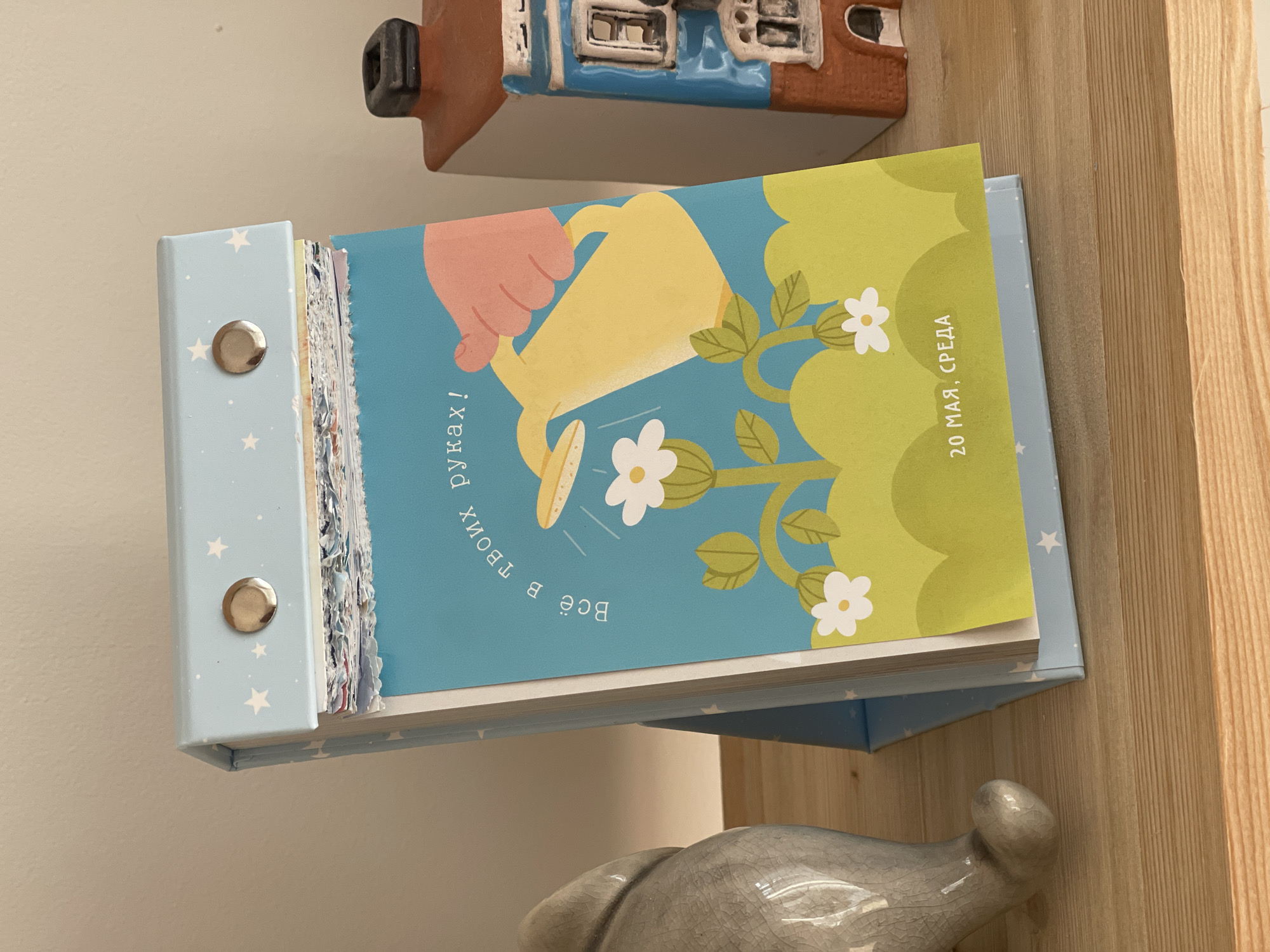 The image shows a calendar propped open. The open page shows a yellow watering can being held by a pink hand, watering a flowering plant. The date "20 мая, среда" (May 20, Wednesday) is visible on the right side of the page. The calendar has a light blue cover with white stars. It is held together with metal brads. The background includes a decorative house figurine and a gray ceramic object on a wooden shelf. The location in Izhevsk, Russia is not discernible from the image.