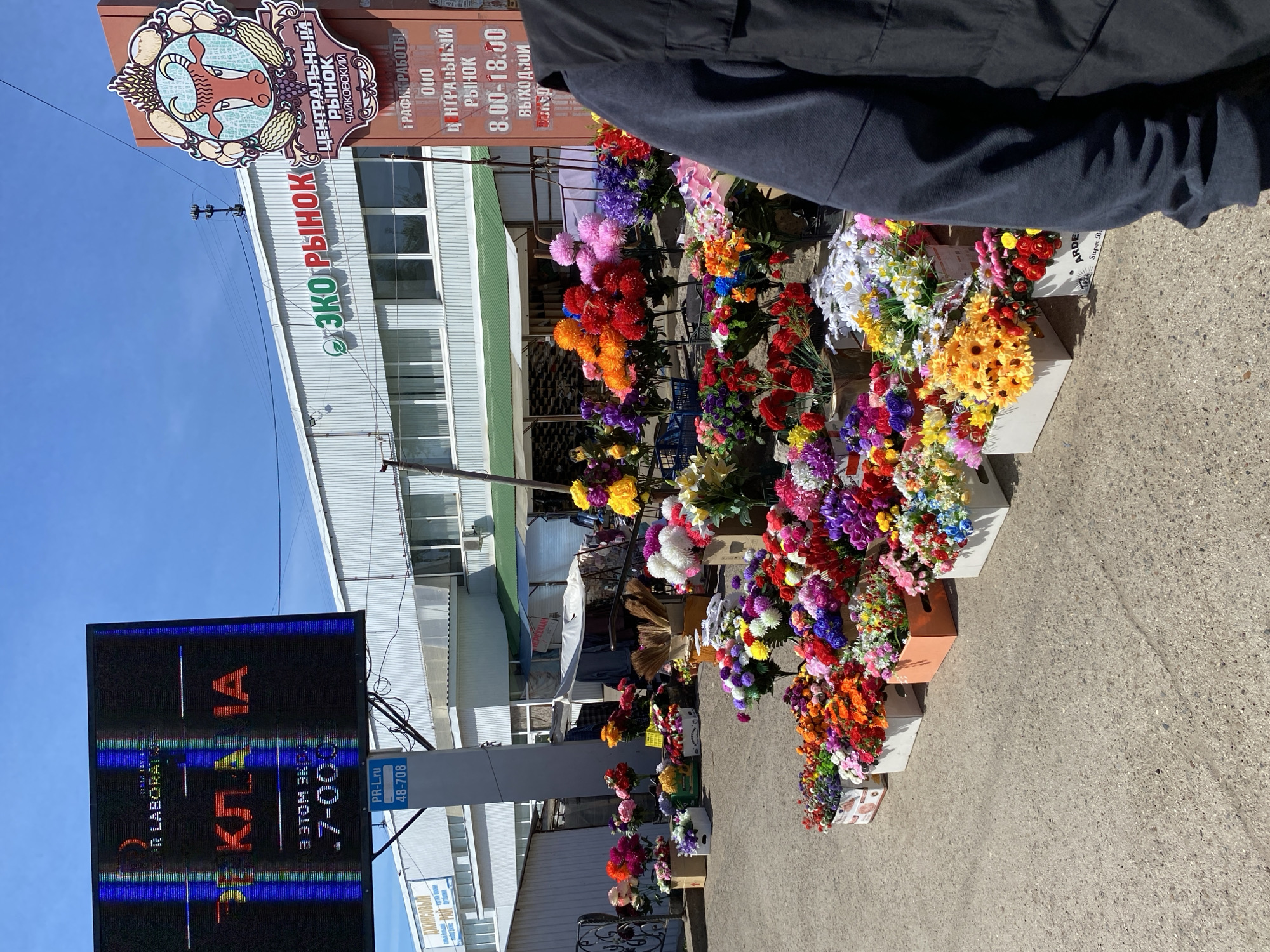 The image depicts a street scene in Chaikovsky, Russia, featuring a flower stall outside the "Central Market." The stall displays a variety of colorful artificial flowers arranged in boxes. Signs are visible for "ECO Market" and a digital display showing the word "KLANA," a phone number, and a URL. A person wearing a dark jacket is partially visible in the upper right of the frame. The overall environment suggests a commercial area within the city.