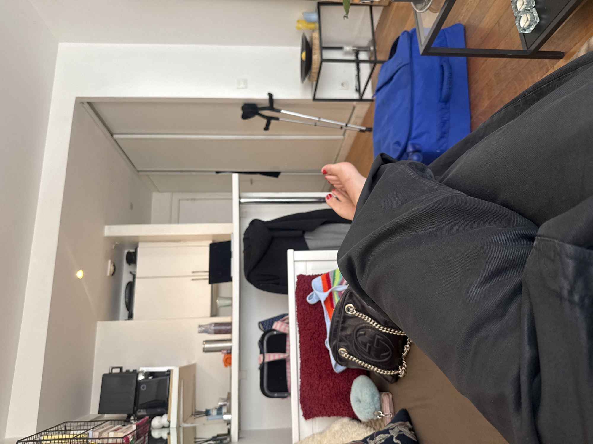 The scene captures an indoor residential space, identified by the prompt as being in Paris, France, from the perspective of a person relaxing with their legs extended. The lower legs and bare feet of a person, wearing black pants and with red nail polish on their toenails, are visible in the foreground. They are resting on a brown surface, likely a couch or bed. Next to the person's legs, a black Gucci handbag with a silver chain strap, a textured red throw pillow, a light blue fluffy object, and a multi-colored striped knit item are present.
The background reveals an open-plan living area and kitchen. To the left, a white kitchen counter and upper cabinets are visible, with various items on the counter and a shelf above it, including a metal thermos, a glass bottle, a cup, and a laptop. A black hooded garment is draped over a chair or similar object near the counter. Further left, a wall-mounted television or monitor and a microwave are stacked on a white shelf. To the right, a pair of metallic crutches with black handgrips and arm cuffs leans against a white wall. Below the crutches, a royal blue duffel bag or piece of luggage rests on the light wood floor. Adjacent to the crutches, a small black metal side table with a glass top holds a dark bowl containing a single yellow fruit, possibly a lemon, and two yellow cylindrical objects. On the lower shelf of the side table, two small glass candle holders each contain a white candle. White sliding doors, possibly for a closet or an adjacent room, are visible in the mid-background. The walls throughout the visible space are white.