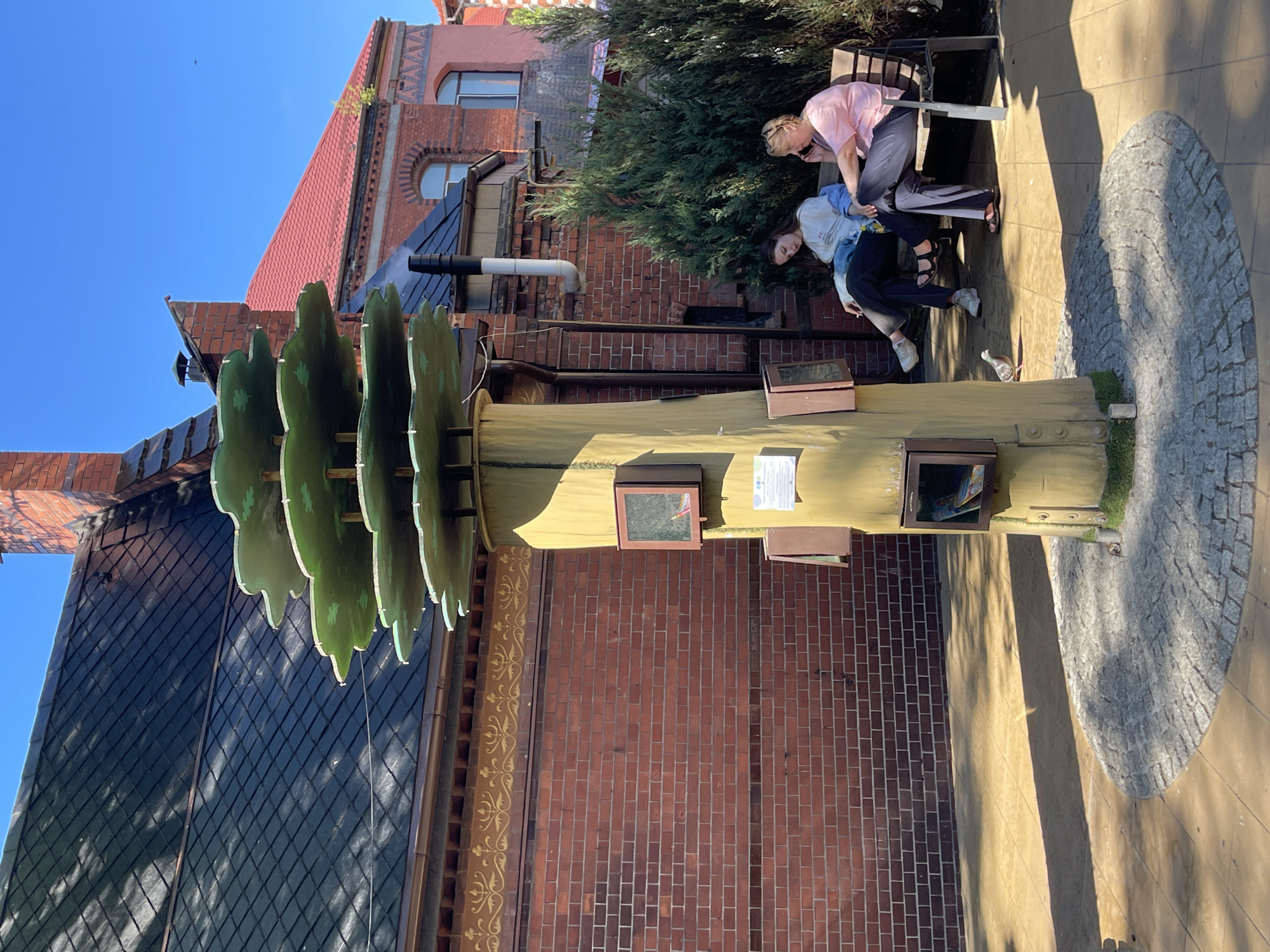 In Zelenogradsk, Russia, a whimsical tree-shaped structure, possibly a public book exchange, stands on a paved area. The structure is yellow and has several square openings with glass fronts displaying small items. Atop the tree are layered, green, leaf-like platforms. In the background, a brick building with a dark tiled roof and red tiled roof is visible, along with a chimney. To the right of the tree, two women are seated on a bench. One woman, on the left, wears a light-colored top and dark pants, and appears to be looking down. The other woman, on the right, wears a pink shirt and dark pants, and is talking on her phone. A pigeon is walking on the pavement in front of the tree. The scene is brightly lit by the sun, casting shadows on the ground.