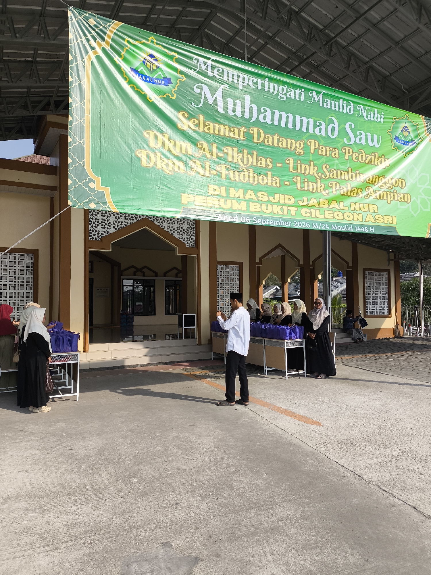 A banner hangs above the entrance to a building, proclaiming "Memperingati Maulid Nabi Muhammad Saw" (Commemorating the Birthday of Prophet Muhammad SAW) and "Selamat Datang Para Pedziki" (Welcome, Visitors). The banner also lists locations and dates: "DKM Al-Ikhlas - Link. Sambiranggon, Dkm Al-Fudhola - Link. Palas Ampian". Below this, it states "DI MASJID JABAL NUR Perum Bukit Cilegon Asri" (At Jabal Nur Mosque, Bukit Cilegon Asri Housing Complex). Further text indicates the date as "Ahad 06 September 2026 M/24 Maulid 1448 H" (Sunday, September 6, 2026 CE/Maulid 24, 1448 AH).
In front of the mosque entrance, several people are gathered. On the left, two women wearing hijabs are standing near tables covered with blue bags. A man in a white shirt and black pants is standing in the center, facing the tables and holding a phone. To the right of the man, a group of women in hijabs are also positioned near the tables. A man in a blue shirt is seated on steps to the right, in the background. The mosque has a beige and brown facade with patterned windows. The ground in front of the building is paved concrete. The scene appears to be an outdoor event at a mosque in the Bukit Cilegon Asri housing complex in Cilegon, Indonesia.