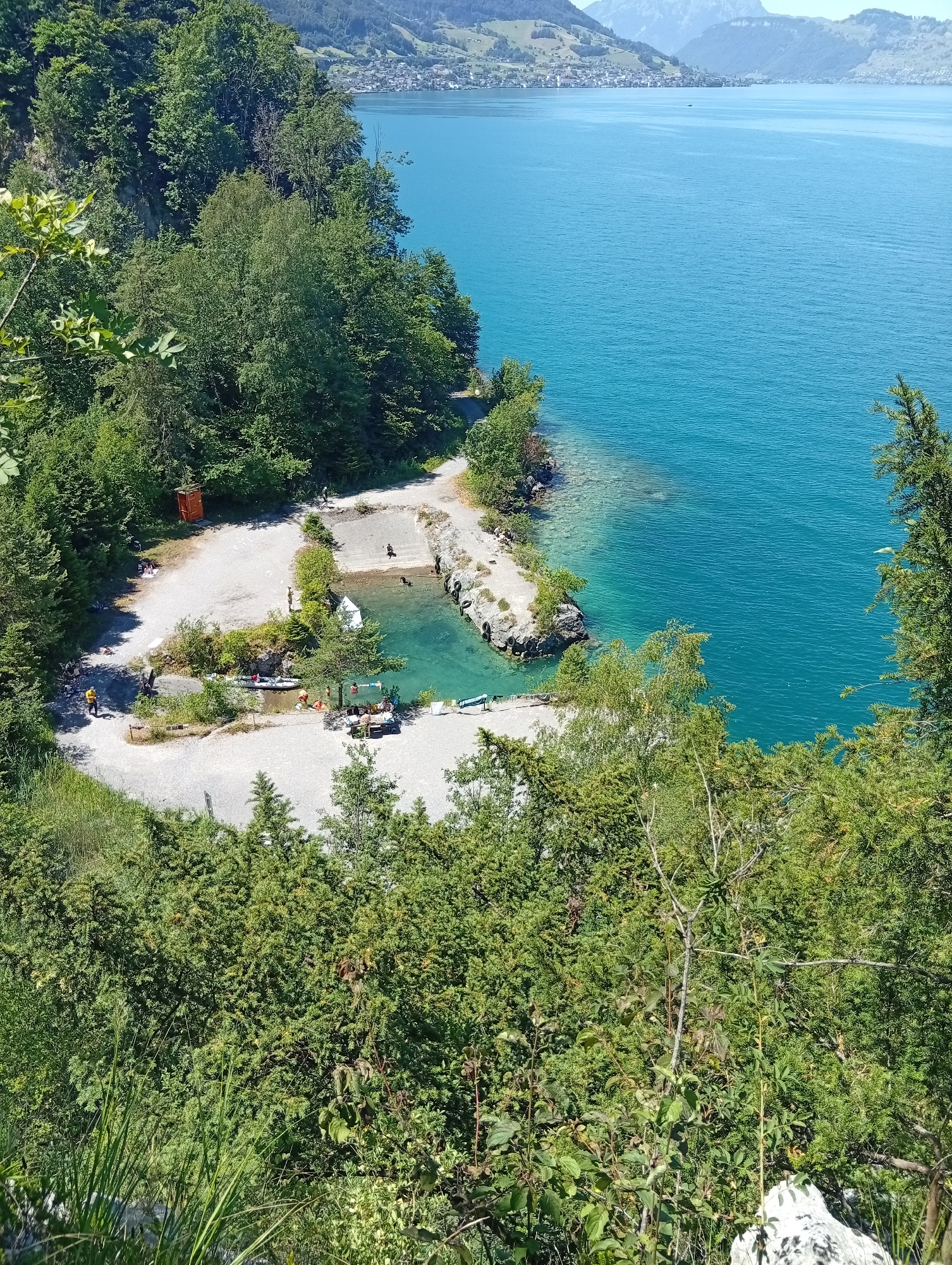 An aerial, high-angle shot captures a small, man-made swimming area carved into the shore of a large, bright blue lake, likely Lake Lucerne, in Emmetten, Switzerland. Lush green trees dominate the foreground and surround the bathing area. The bathing area is a rectangular pool with a concrete base and natural rock formations along one side. Clear, turquoise water fills the pool. Several people are visible in and around the water. Some are swimming, while others are relaxing on the gravelly beach.
A small white raft or boat is anchored in the pool, and a few inflatable tubes are scattered on the beach. Nearby, a small, red wooden structure, possibly a changing room or outhouse, stands on the sandy area. A few other individuals are scattered on the gravel paths and the beach, some appearing to be walking or sitting. A canoe or kayak is pulled up onto the shore, next to some life vests. The main body of the lake is expansive and a vibrant blue, with distant green hills and mountains framing the horizon. The overall impression is a popular, serene recreational spot on a sunny day.