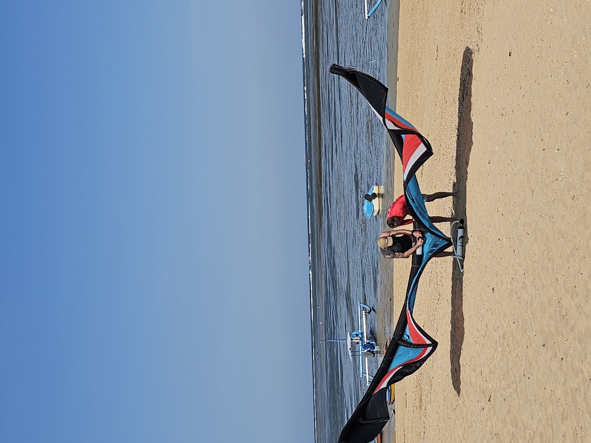 Two individuals are on a sandy beach near the water's edge, preparing a large, colorful kite for use. The kite, predominantly blue, red, and black, is laid out horizontally, with its wings partially inflated. One person, wearing a tan baseball cap and a dark sleeveless shirt, is bending over the kite, appearing to adjust it. The second person, wearing a red shirt, is also focused on the kite, with their hands engaged with it.
The beach is composed of light brown sand with scattered small shells. In the background, the ocean is visible, with shallow, calm water and a gentle wave line on the horizon. A traditional Indonesian outrigger boat, painted blue, is situated in the water. Another smaller, light-colored boat is also present further out. The sky is clear and bright blue, indicating a sunny day. The scene is likely from a beach in or near Denpasar, Indonesia, given the characteristic outrigger boats and the clear, tropical coastal environment.