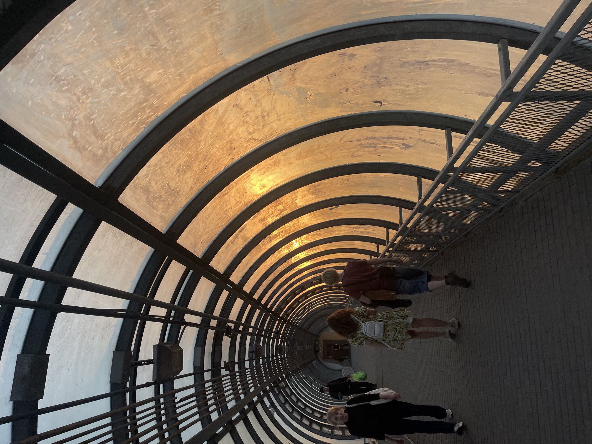 Captured from a low angle, the image depicts an enclosed pedestrian bridge, likely located in Khimki, Russia. The bridge has a semicircular transparent roof supported by multiple metal arches. Natural light streams through the roof, creating a warm, golden glow within the tunnel.
The bridge is occupied by four people walking along its length. The individuals appear to be of varying ages and are dressed in casual attire. Their actions consist of walking. The environment is clean and appears well-maintained, with a brick or concrete walkway. A metal grate walkway runs along the top right side of the image.