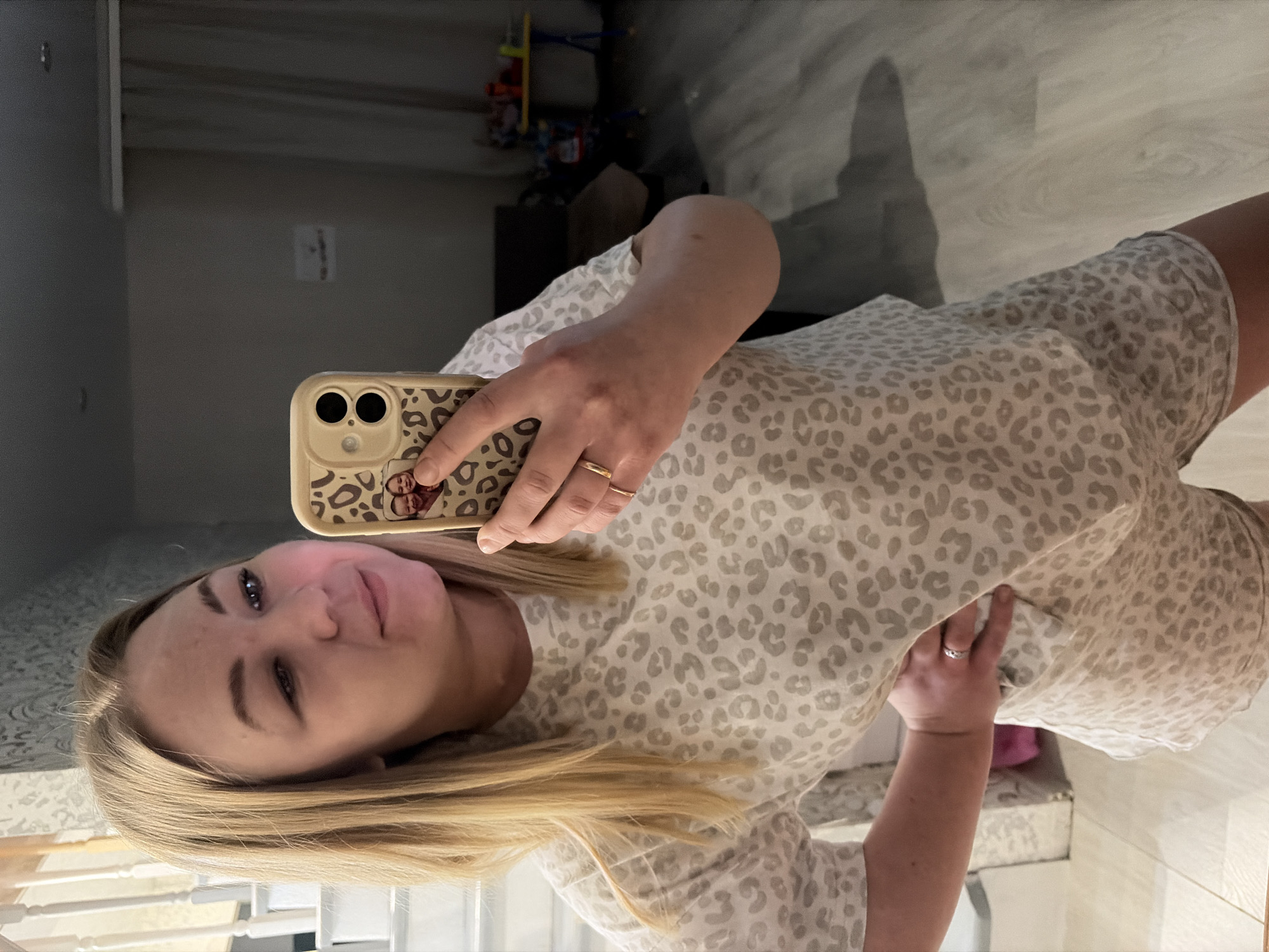 The image displays a woman taking a mirror selfie indoors, likely within a home setting in Noviy, Russia. The woman, who has long blonde hair, fair skin, and light-colored eyes, looks towards the camera with a neutral to slightly smiling expression. She is dressed in a casual two-piece pajama set featuring a light grey and beige leopard print pattern, consisting of a short-sleeved top and matching shorts.
She holds a light tan-colored smartphone with a leopard print case in her left hand. The phone case clearly shows two camera lenses on its back, and a small, blurred image depicting two faces (possibly children or a couple) is visible on the case near her index finger. A gold ring adorns the ring finger of her left hand. Her right hand is gently placed on her stomach or side, and a silver-colored ring is visible on its ring finger.
The background reveals a light-colored wooden floor and a plain grey wall with some indistinct marks. A reflection in the mirror suggests a railing or staircase in the deeper background, and some blurry objects, potentially toys or furniture, are visible further away. The indoor lighting creates a general bright ambiance. No discernible text is visible within the image beyond the blurry photo on the phone case.