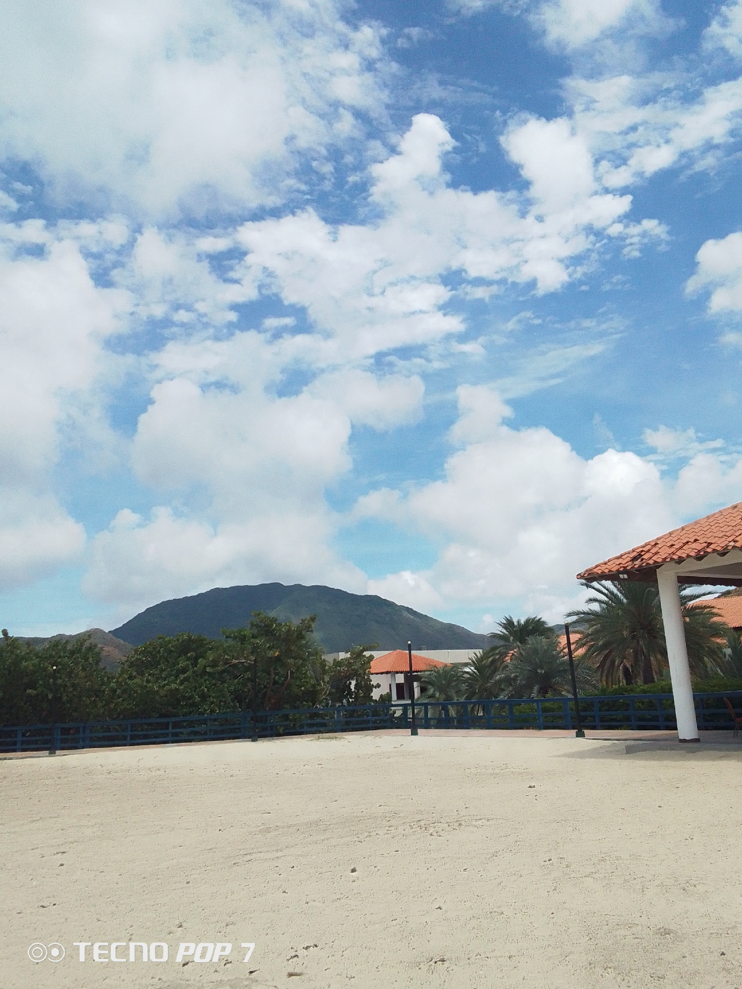 The image shows a view in Pedro González, Venezuela. In the foreground is a large expanse of light-colored sandy ground. A dark blue fence runs across the midground, separating the open area from a line of trees and palm trees. Behind the trees, there are buildings with orange roofs. In the distance, a dark green mountain rises against a blue sky filled with scattered white clouds. On the right side of the frame, a building with an orange tile roof and white columns is visible. The sky occupies a significant portion of the frame, showcasing the cloud formations. The bottom left corner displays the text "TECNO POP 7".