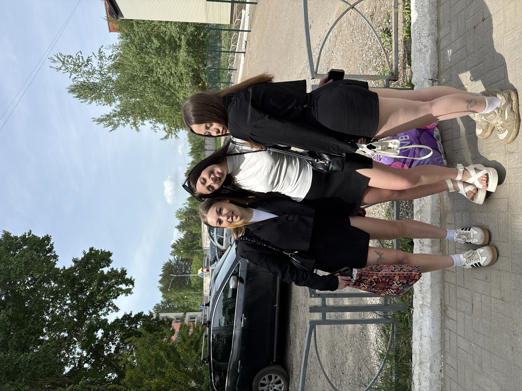 Three young women are posing in an outdoor scene, likely a street in Chaikovsky, Russia, indicated by the Cyrillic script 'NB' on a bag one of them carries. The women are lined up along a sidewalk with a low metal railing behind them and a dark SUV parked nearby. They are all wearing variations of a similar outfit: black blazers, skirts or shorts, and white tops. The woman in the front has blonde highlights in her hair and is holding a patterned bag. The woman in the middle wears sunglasses on top of her head, and the woman in the back is holding a smartphone. Their footwear varies. Trees and buildings are visible in the background under a partly cloudy sky.