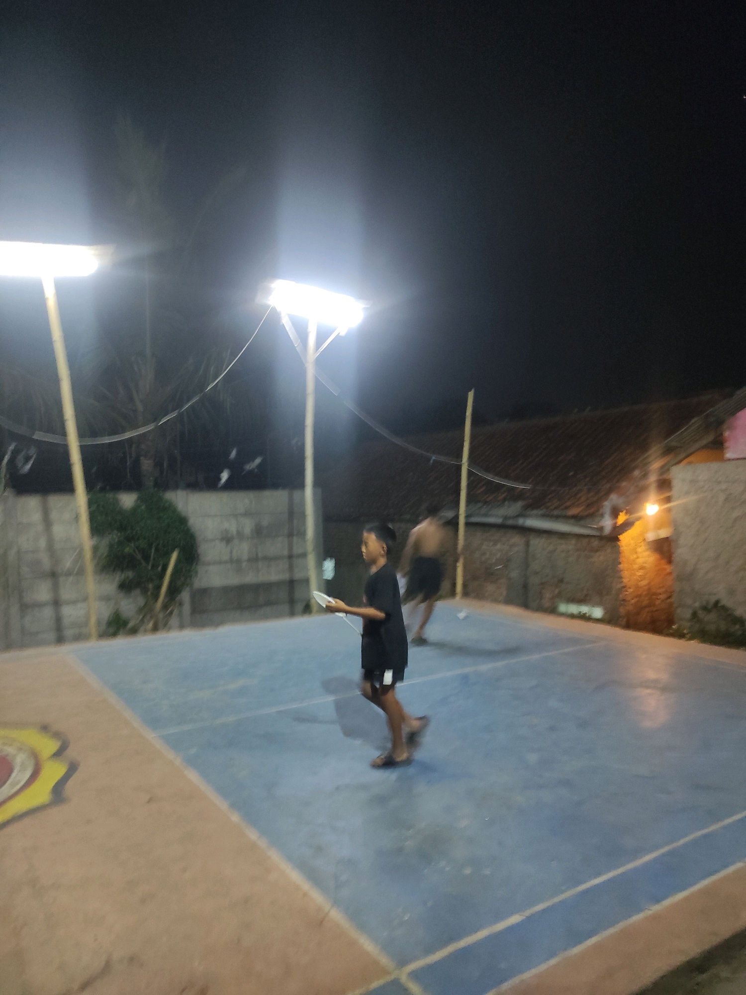 In Padalarang, Indonesia, at night, a group of individuals are on an outdoor court, likely a badminton or volleyball court given the net visible in the background. Two young males are the primary subjects. One, wearing a black shirt and shorts, is in the foreground, holding a white object, possibly a phone or a paddle, and appears to be mid-stride. The other male, shirtless and wearing dark shorts, is in the mid-ground, running or moving rapidly across the court. The court itself is divided into blue and brown sections, with white lines marking the boundaries. Two tall lampposts with bright lights illuminate the scene, casting long shadows and creating a high-contrast environment. In the background, a concrete wall, some foliage, and a building with a tiled roof are visible. The overall impression is of an active recreation scene taking place after dark, facilitated by artificial lighting.