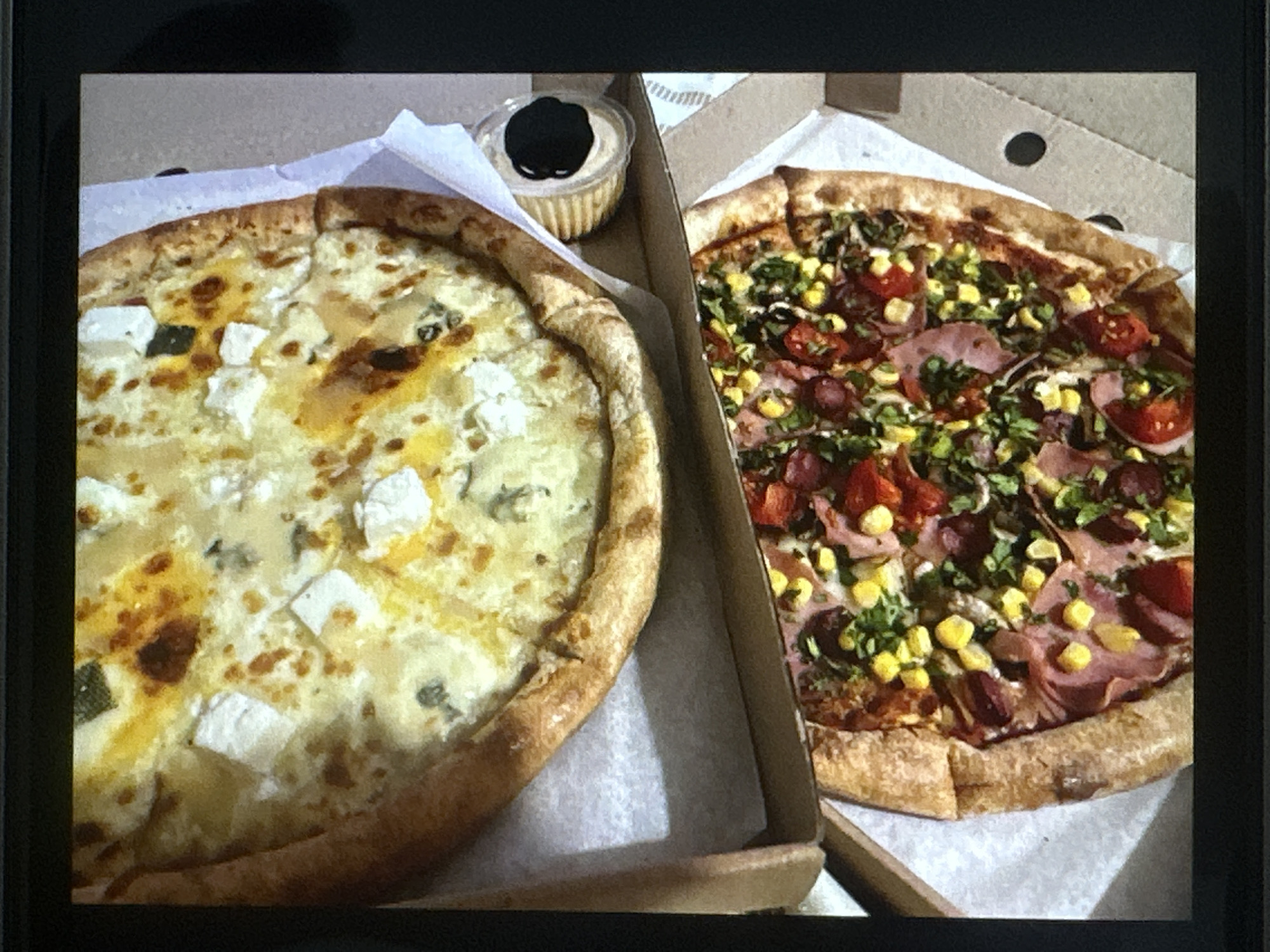 The image shows two pizzas in cardboard pizza boxes. The pizza on the left is topped with a light-colored cheese, possibly mozzarella or ricotta, and has a thick, golden crust. A small plastic container of sauce sits next to it. The pizza on the right has a more diverse array of toppings, including corn, sliced meat (possibly ham or prosciutto), small green leafy vegetables, and red circular slices (likely tomato or pepperoni). Both pizzas appear to be freshly made and are presented in their delivery boxes. The location is unknown.