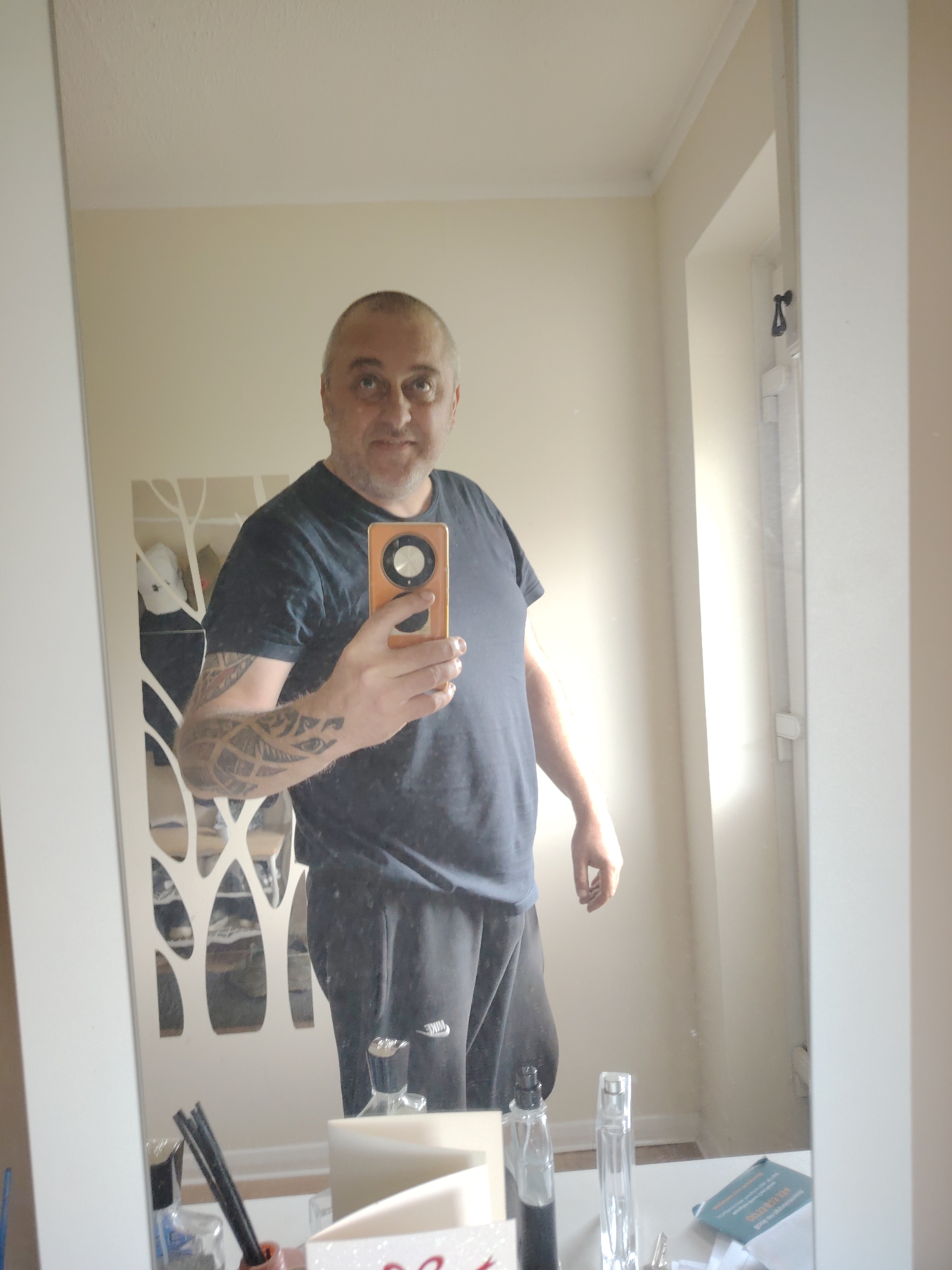 An indoor mirror selfie taken in Huntingdonshire, United Kingdom, shows a middle-aged man with a shaved head and light facial hair, wearing a dark blue short-sleeved t-shirt and dark sweatpants with a white "NIKE" logo on the left thigh. He is holding an orange-cased smartphone with a large circular camera module in his right hand, reflecting his image in a large white-framed mirror. Tribal or geometric tattoos are visible on his right forearm. Behind his reflection, a decorative white wall panel with a cutout tree or organic pattern is visible on the left, with vague reflections of items behind it. The room features light beige walls and a white ceiling with coving. To the right, a window or doorway with a white frame is visible, admitting bright natural light. The bottom of the mirror reflects a white surface (likely a dresser or shelf) on which various objects are placed: several bottles (including clear glass perfume bottles and a small blue-liquid bottle), a red-orange holder containing black diffuser sticks, a small blue booklet or card, and faintly visible keys.