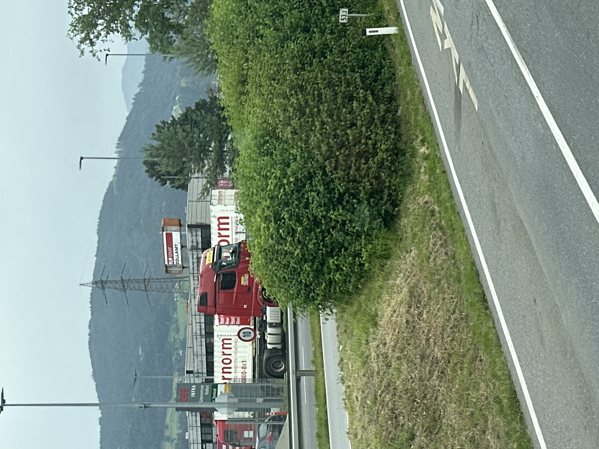 The image depicts a red semi-truck with a white container in Wolfurt, Austria. The truck is parked near a customs building and a facility labeled "INTERNORM." The background shows a landscape with hills, trees, and power lines. The foreground features a road with white markings and a grassy embankment. A small white sign with "53.2" is visible on the right side of the image. The sky is overcast.