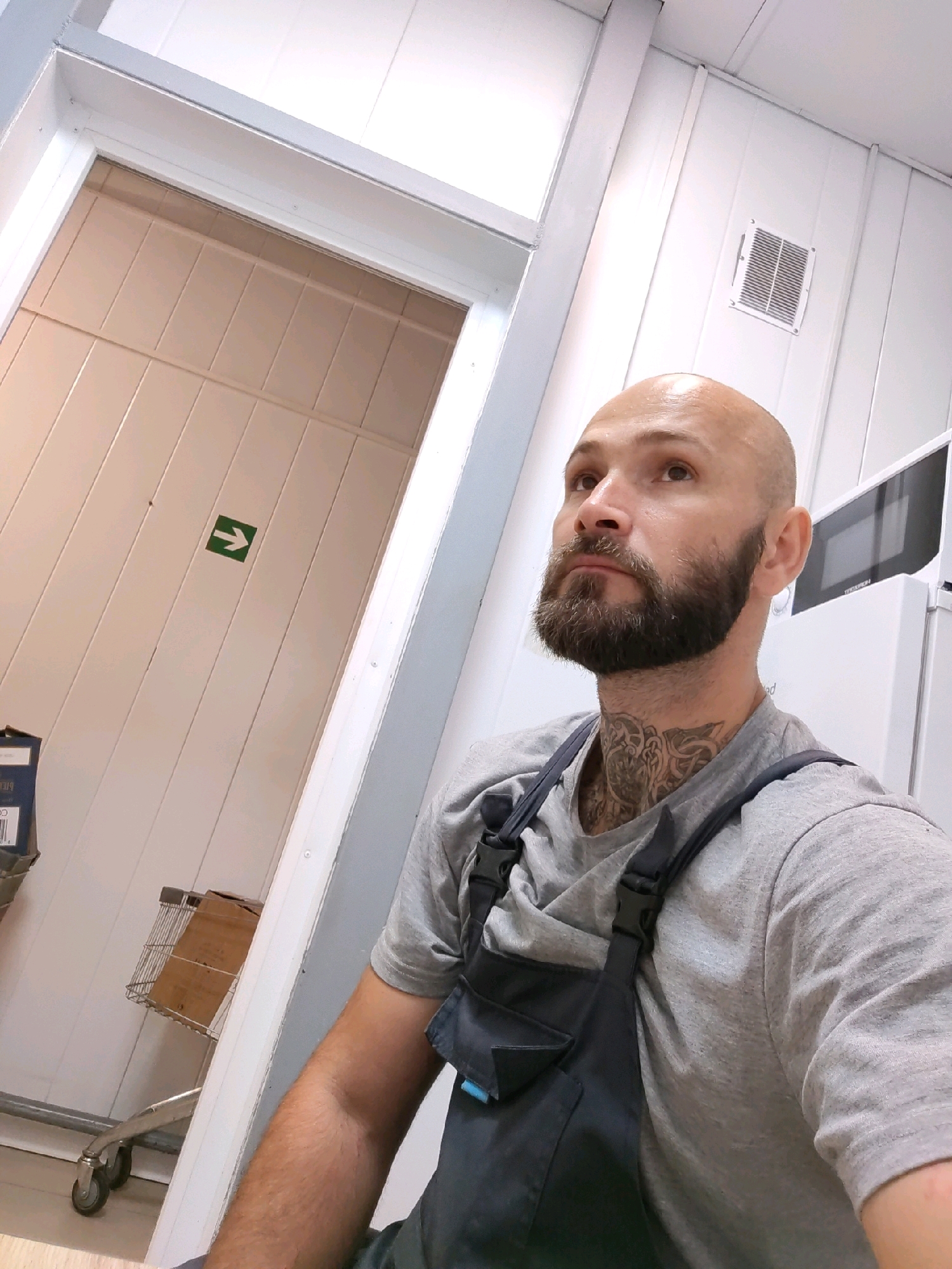 This is a selfie of a bald, bearded man wearing a gray t-shirt and dark blue overalls. He has a tattoo on his neck. The man is standing indoors, in what appears to be a storage or utility area. Behind him, on the left, is a doorway with light-colored paneling and a green arrow sign pointing right. To the right of the doorway is a metal cart with a cardboard box on it. The walls are white paneled, and there is a white vent on the right wall. A microwave is visible on the far right. The image does not provide any specific details that would allow for an accurate determination of the location within the city of Pinsk, Belarus.