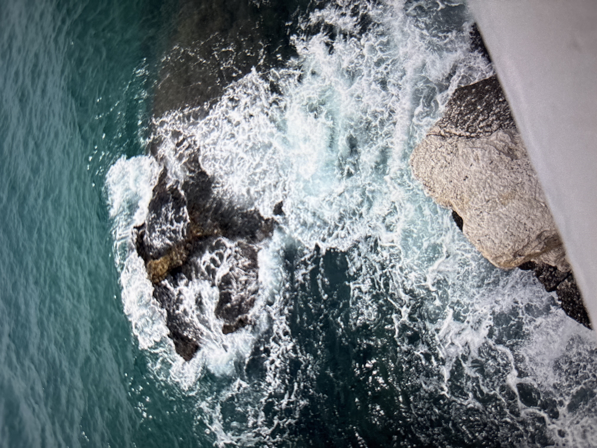 An aerial view captures the turbulent sea crashing against dark, rugged rocks. The water, a deep teal color, is a dynamic expanse of churning white foam and darker currents, highlighting the force of the waves. In the foreground, a large, light-colored rock with a rough texture juts into the frame, its surface appearing weathered and uneven. The scene is devoid of human presence and structures, focusing solely on the raw power of nature where the sea meets the land. The image does not depict any recognizable landmarks or features of Sofia, Bulgaria.