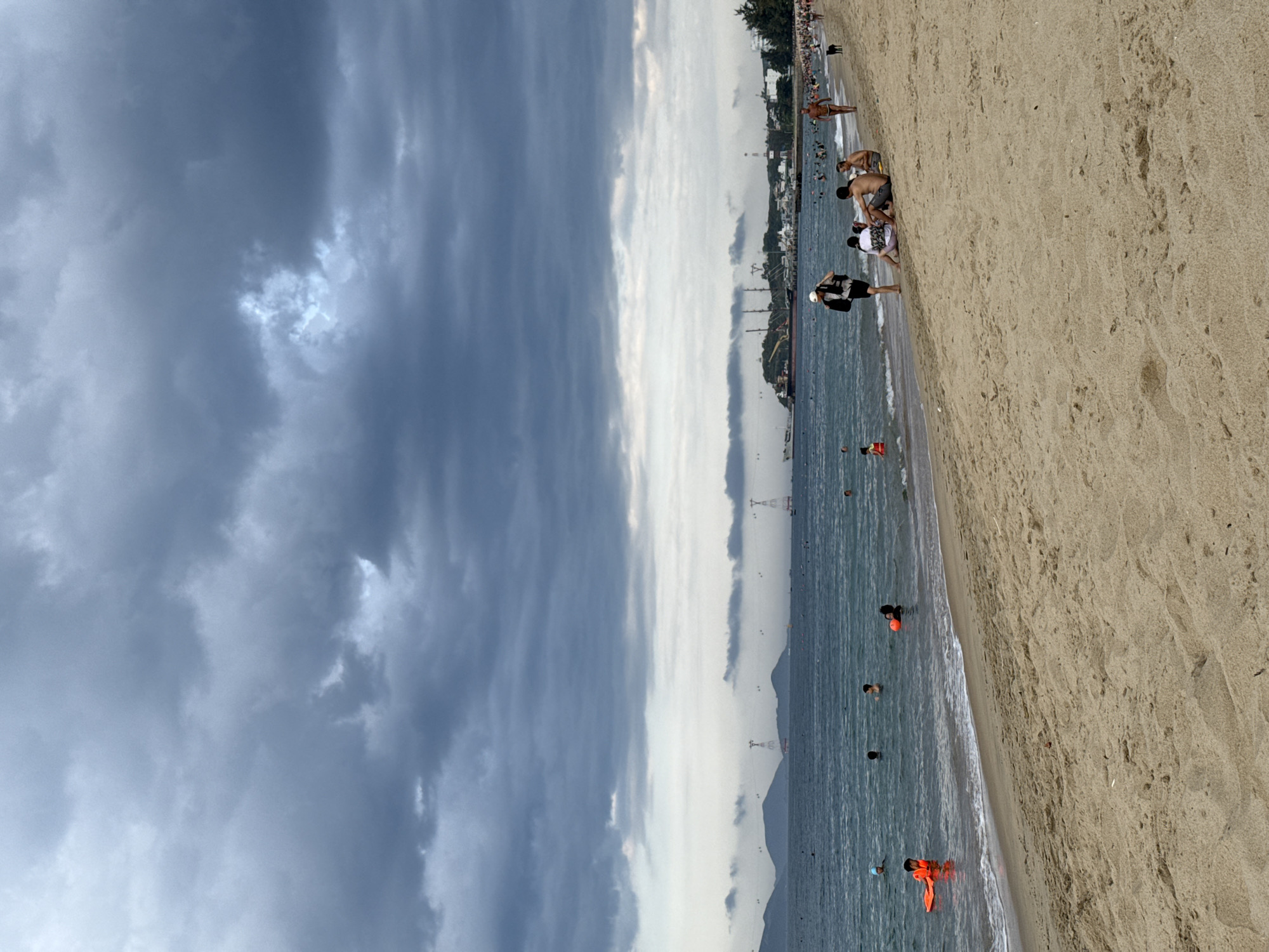 The image depicts a cloudy day at a beach in Nha Trang, Vietnam. The foreground is dominated by a sandy beach with a few people sitting and walking. In the middle ground, the turquoise ocean is filled with swimmers and individuals in orange life vests. The background reveals a coastline with mountains, a harbor with boats and ships, and a city skyline. The overall atmosphere is one of leisure and outdoor recreation under a dramatic, overcast sky.