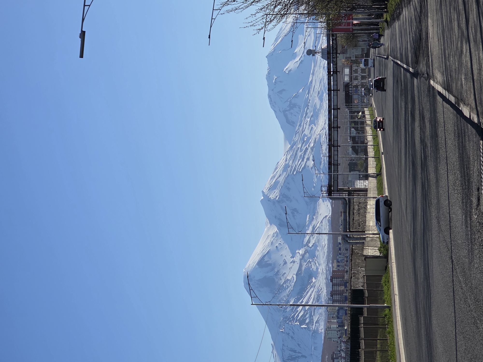 Here is a description of the image:
The image shows a street scene in Petropavlovsk-Kamchatsky, Russia, with a mountain range in the background. The street has a dark asphalt surface and a concrete sidewalk. Several light posts line the street, positioned between the road and the sidewalk. A few buildings are visible. Cars are driving on the street. Several individuals are present along the road, walking. The mountains in the background are covered in snow. The sky above is clear and blue.